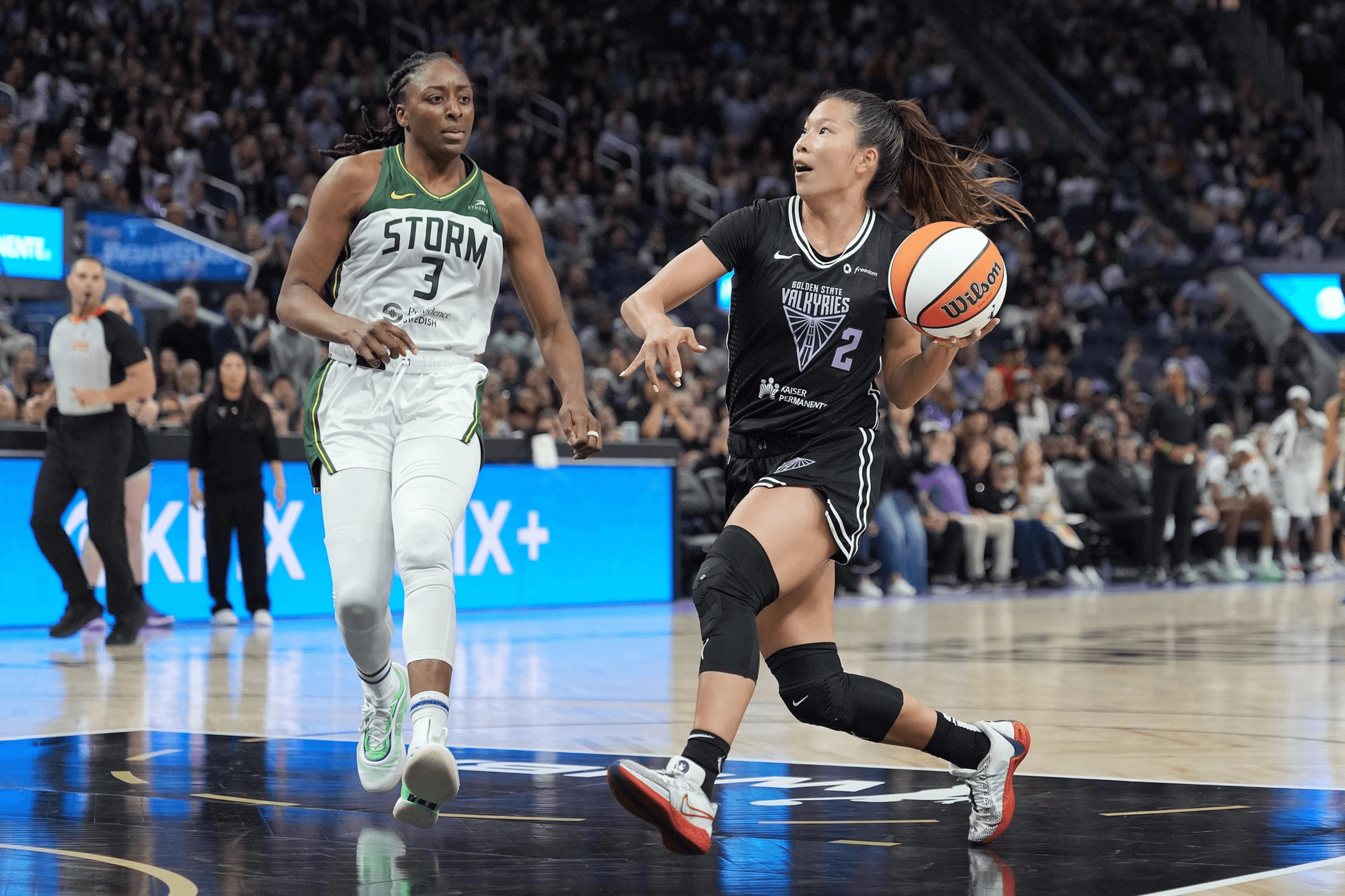 A basketball game is in progress with one player in black dribbling the ball as a player in white defends. The crowd is visible in the background.