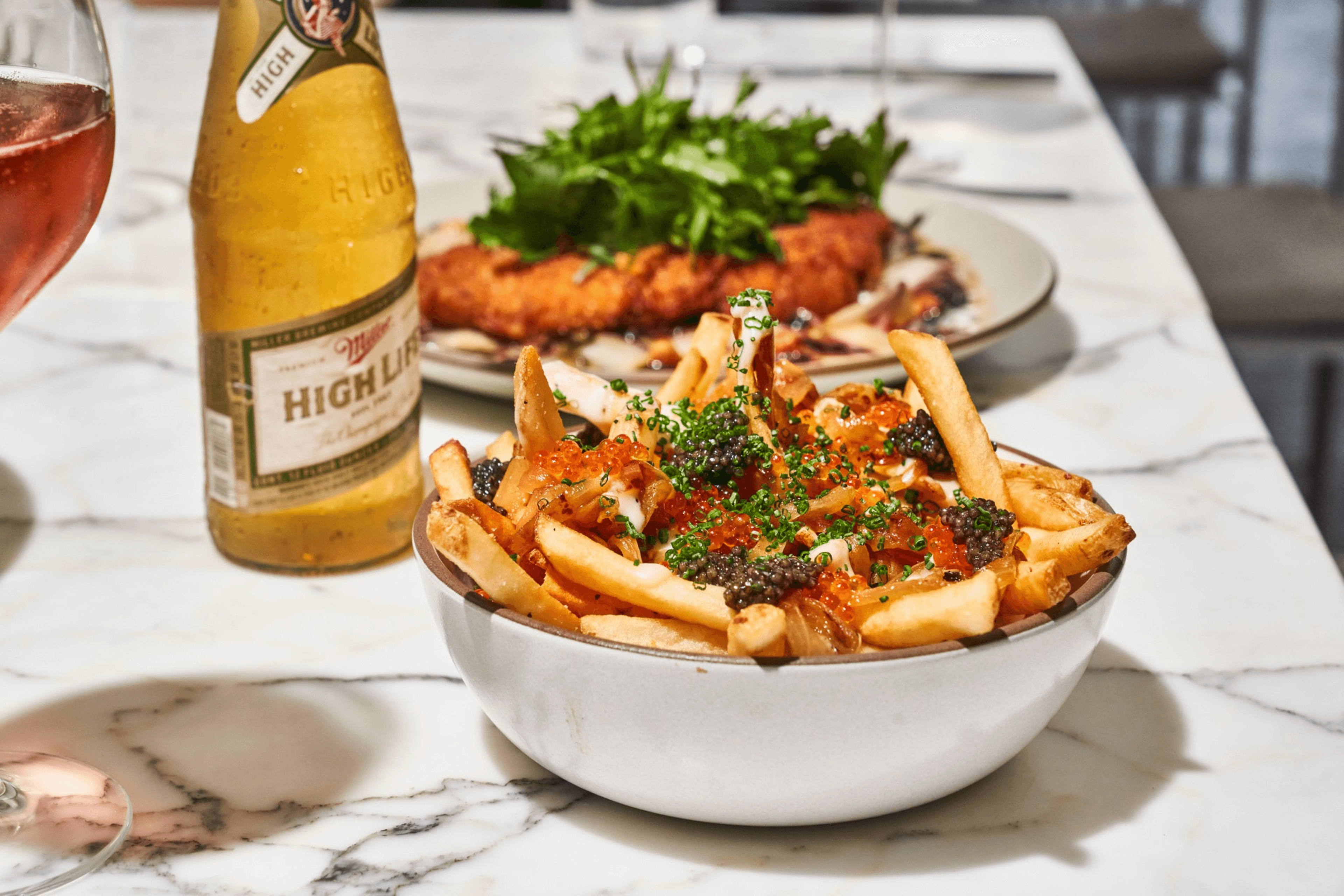A bowl of fries topped with caviar and herbs, accompanied by a bottle of beer. In the background, there's a plate of food with greens.