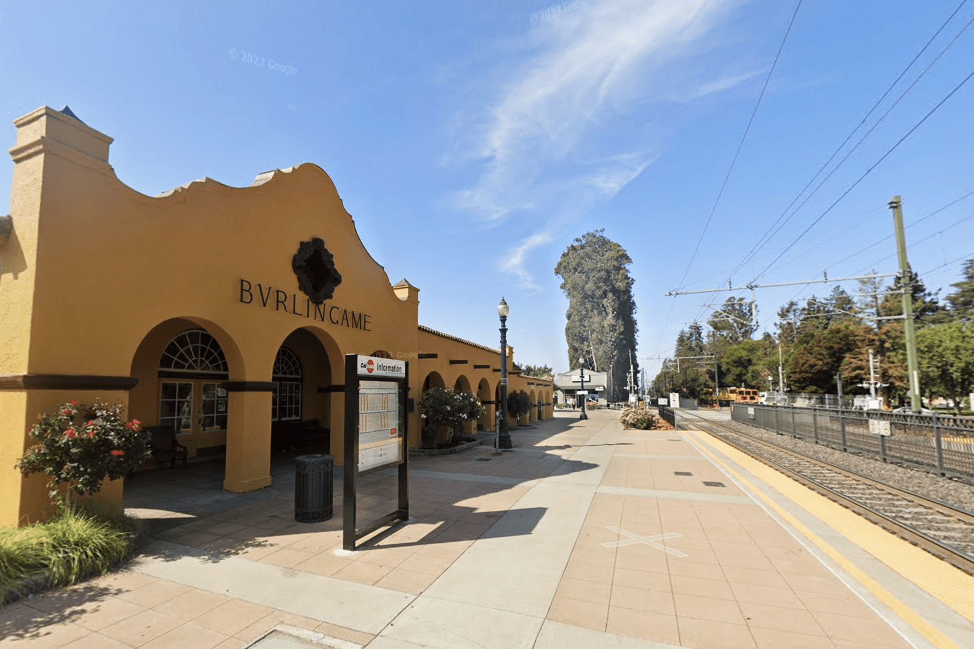 A Spanish-style train station with "BURLINGAME" on the facade, beside a platform and tracks, under a clear sky.