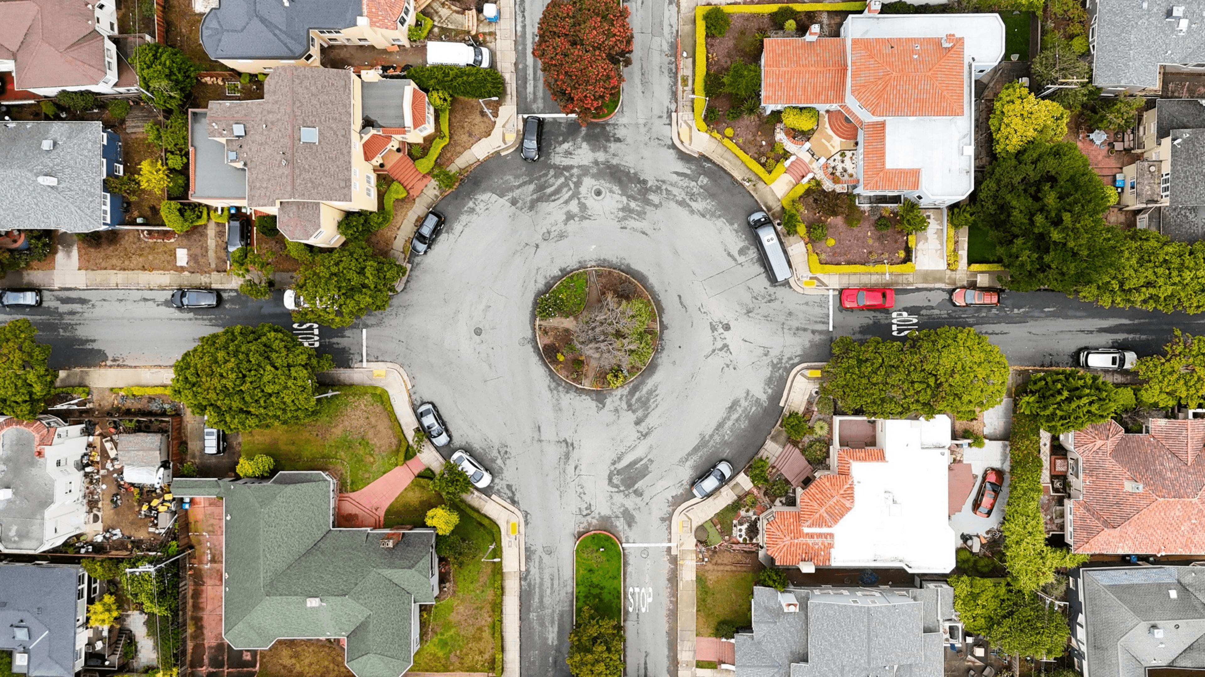 A roundabout intersection with cars parked along the streets and houses surrounding it with trees and gardens nearby.