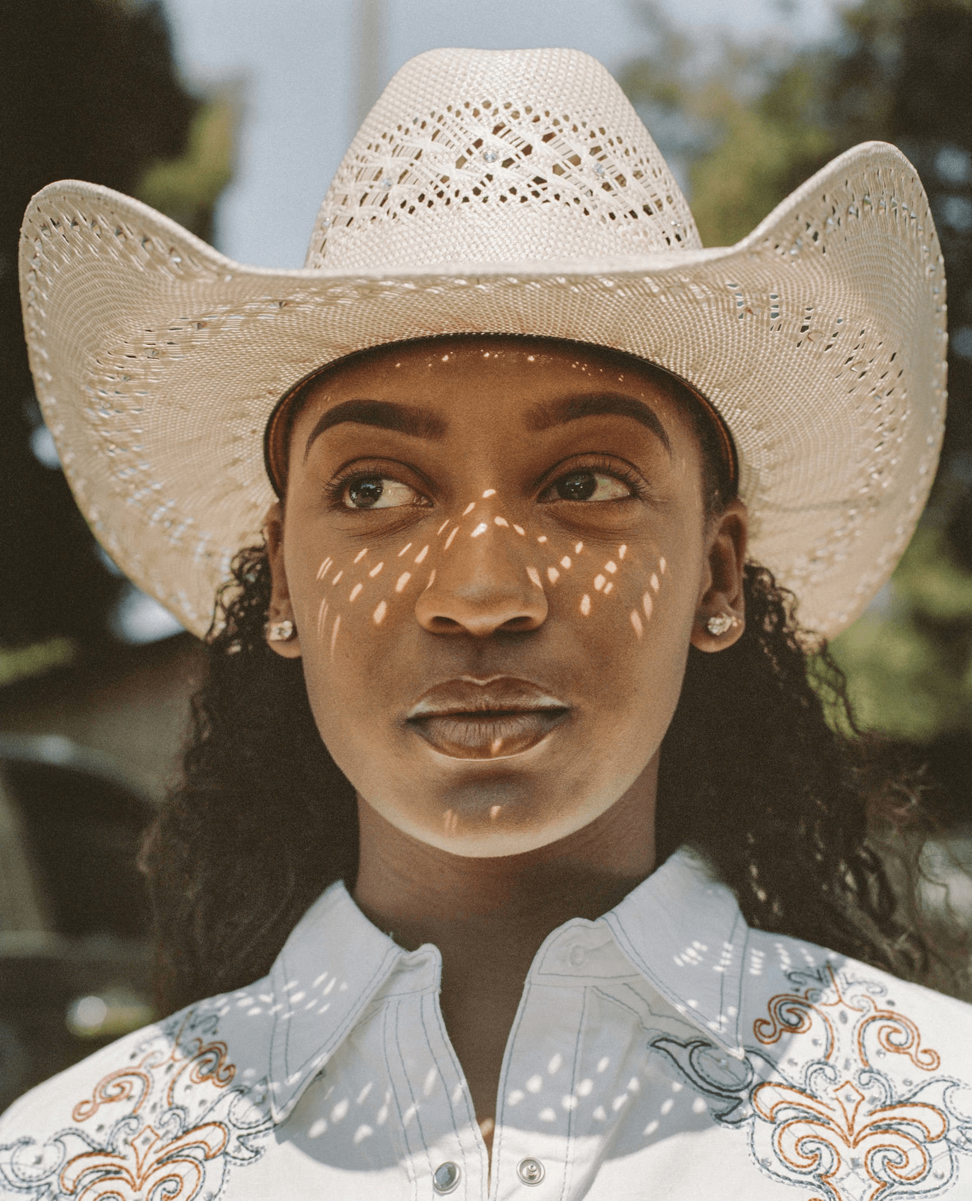 A woman wears a patterned straw cowboy hat casting dotted shadows on her face. Her shirt has colorful embroidery, and she stands outdoors under a clear sky.