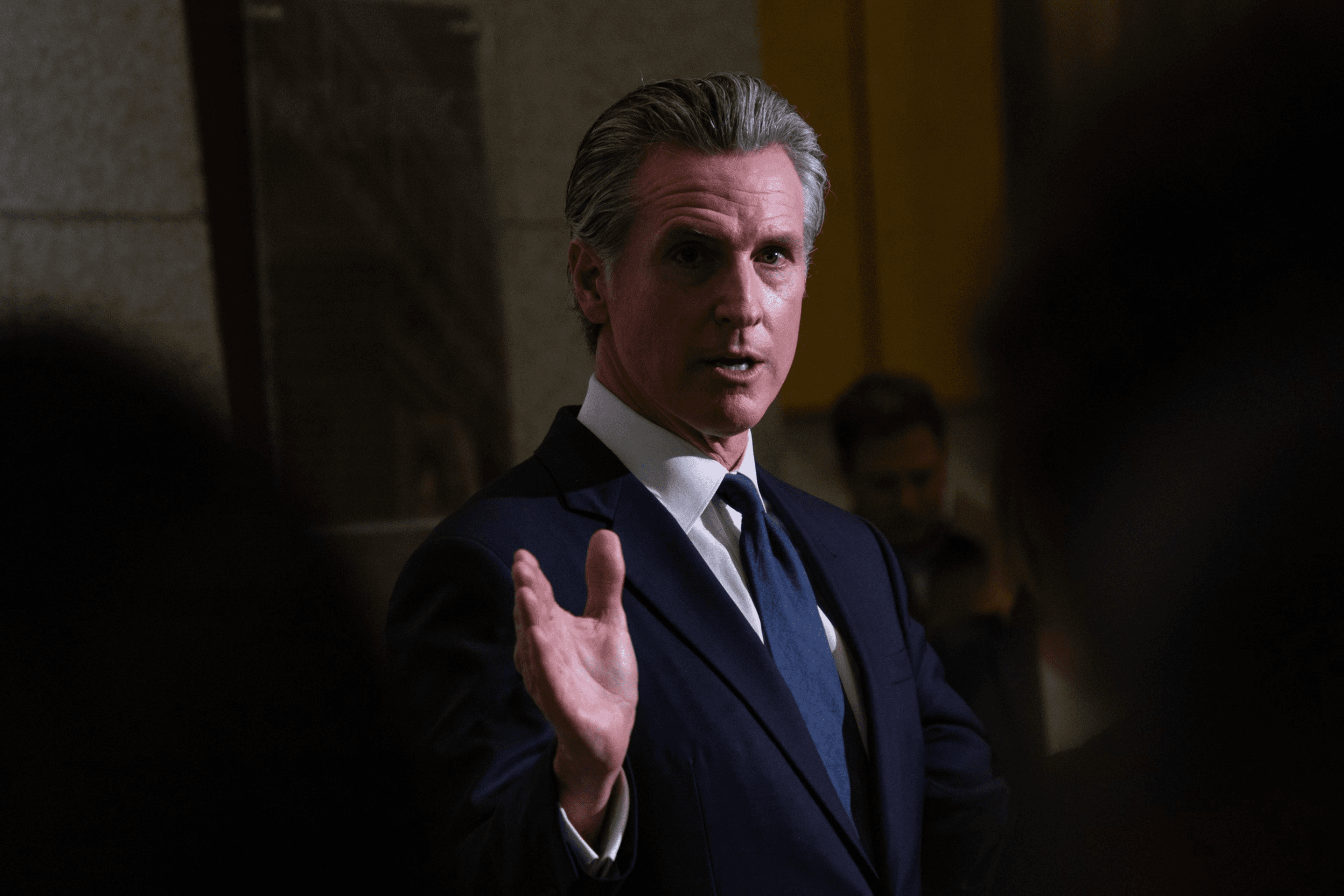 A man in a suit is speaking with an expressive hand gesture. He has short, gray hair and is in a dimly lit indoor setting, suggesting a formal event or meeting.