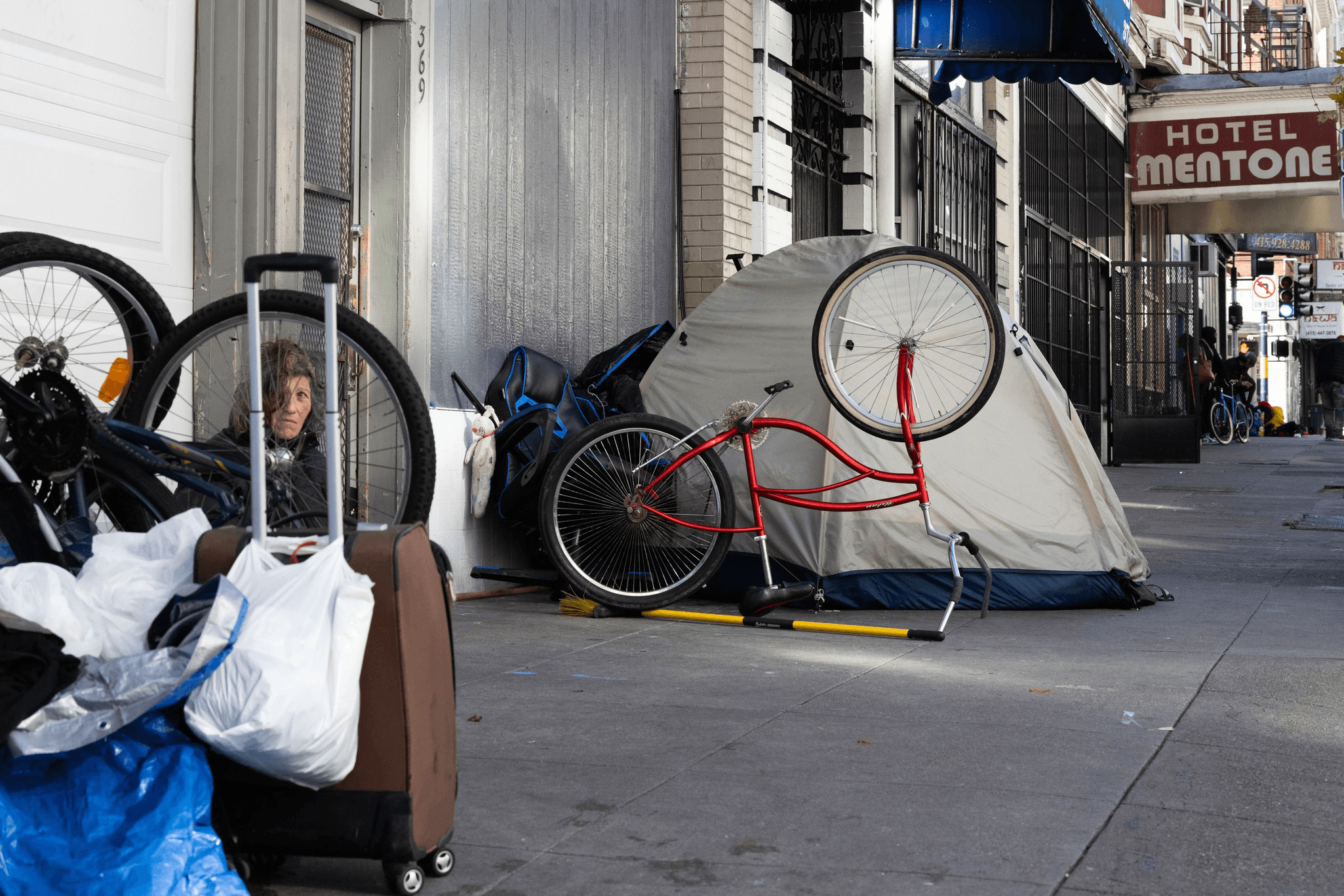 A person sits on a sidewalk beside a tent, surrounded by bicycles, luggage, and bags. A sign for Hotel Mentone is visible above the street.