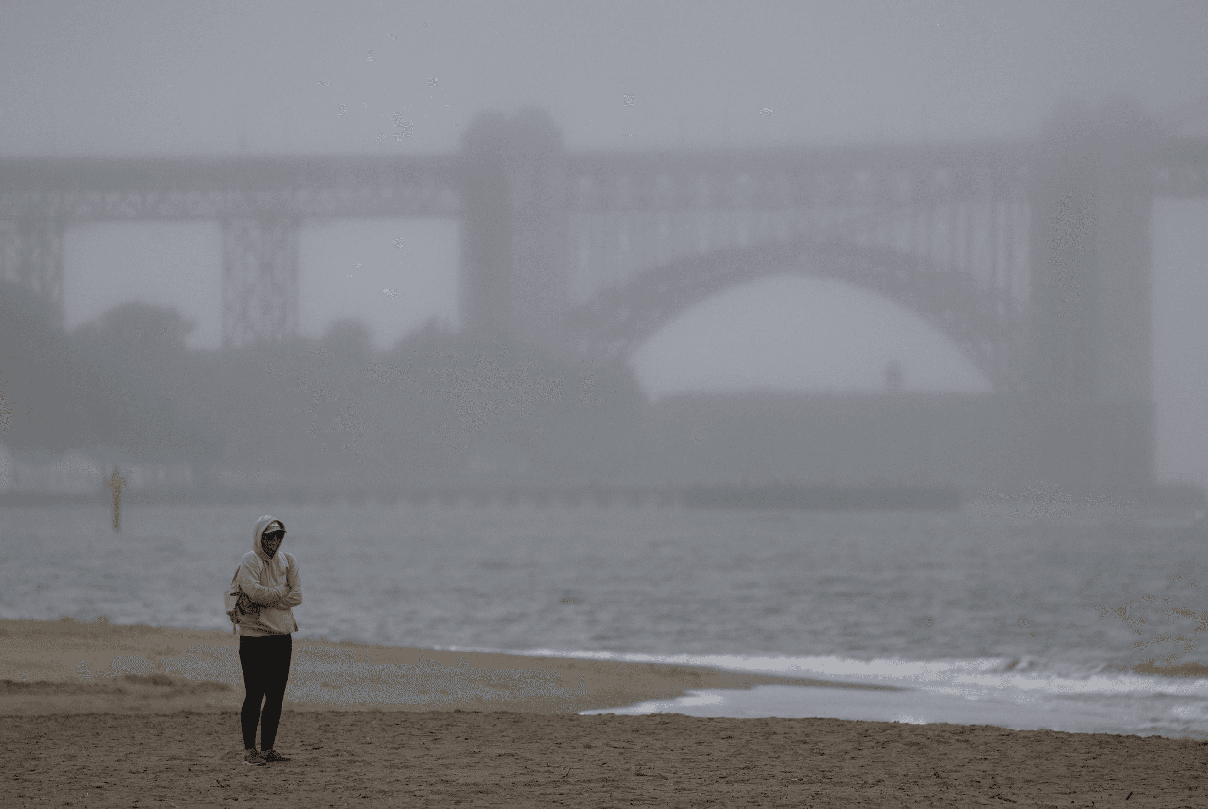 A person in a hoodie walks on a foggy beach near the shoreline. In the background, a large bridge looms faintly in the mist.