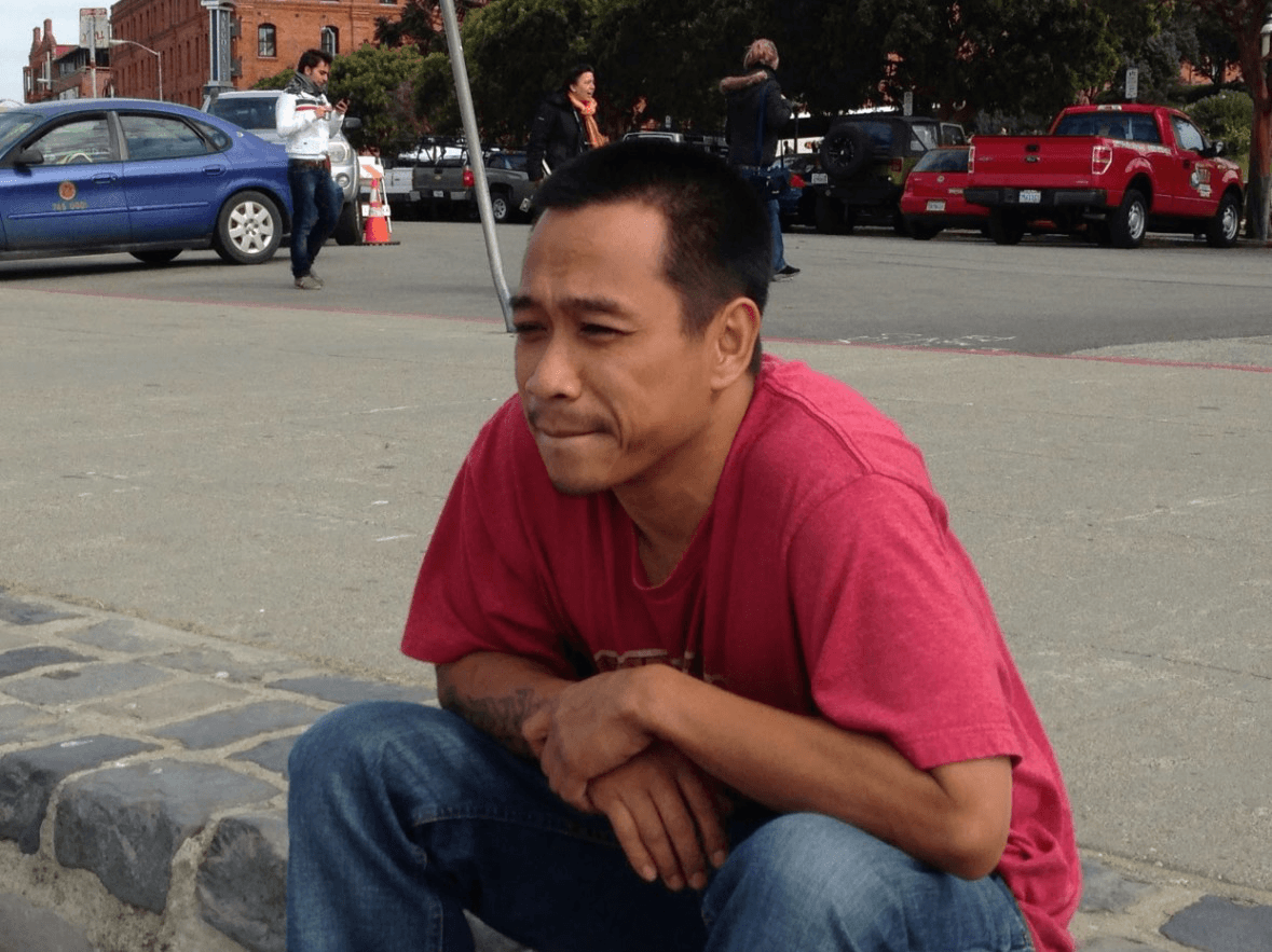 A person in a red T-shirt is crouching on a pavement near parked cars and people. They appear deep in thought, with a street and building in the background.