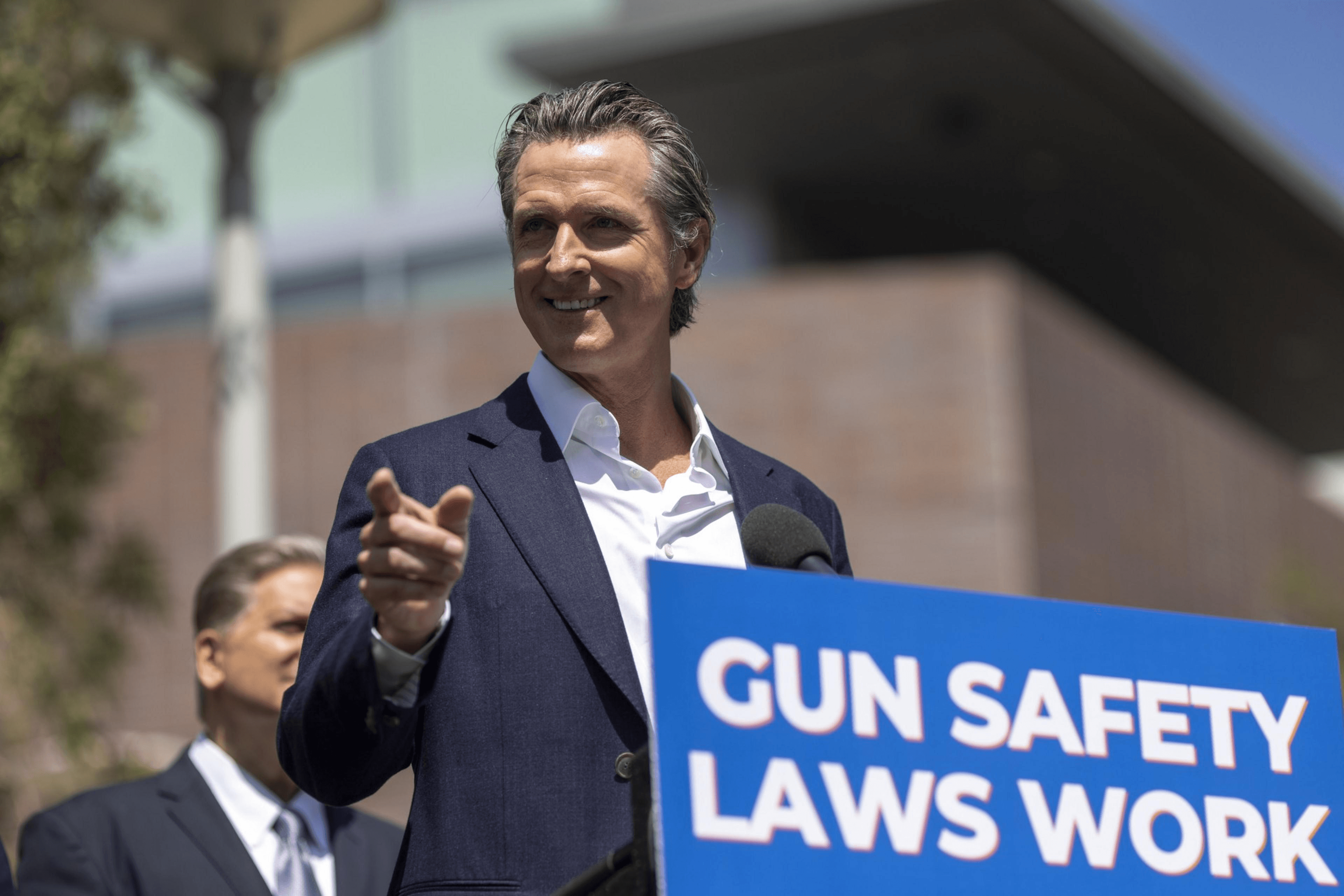 A man in a suit speaks at a podium with a sign reading "Gun Safety Laws Work." Another person stands in the background, and a building is visible behind them.