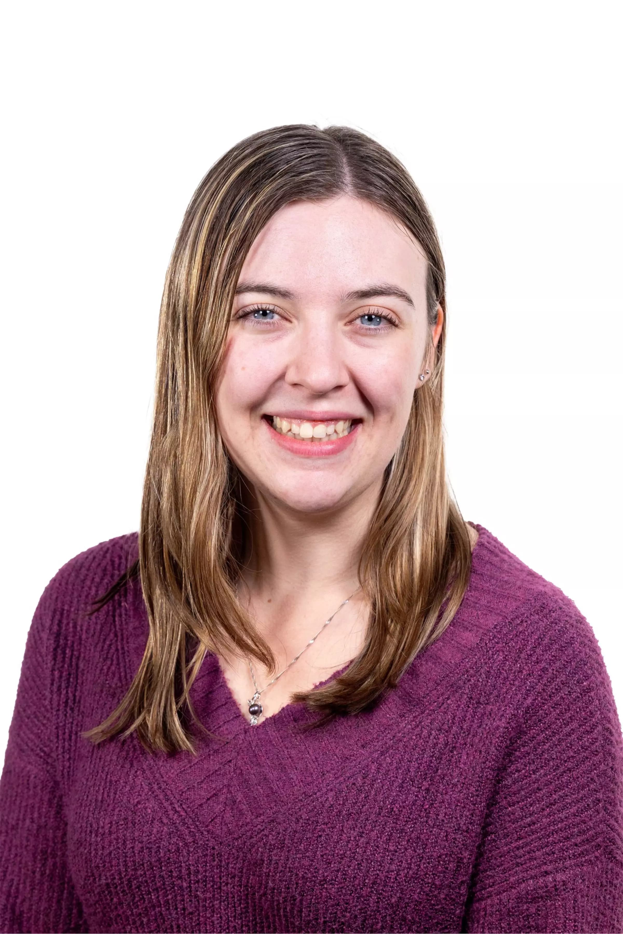 A young woman with straight light brown hair smiles, wearing a purple knit sweater and a delicate necklace with a pendant.