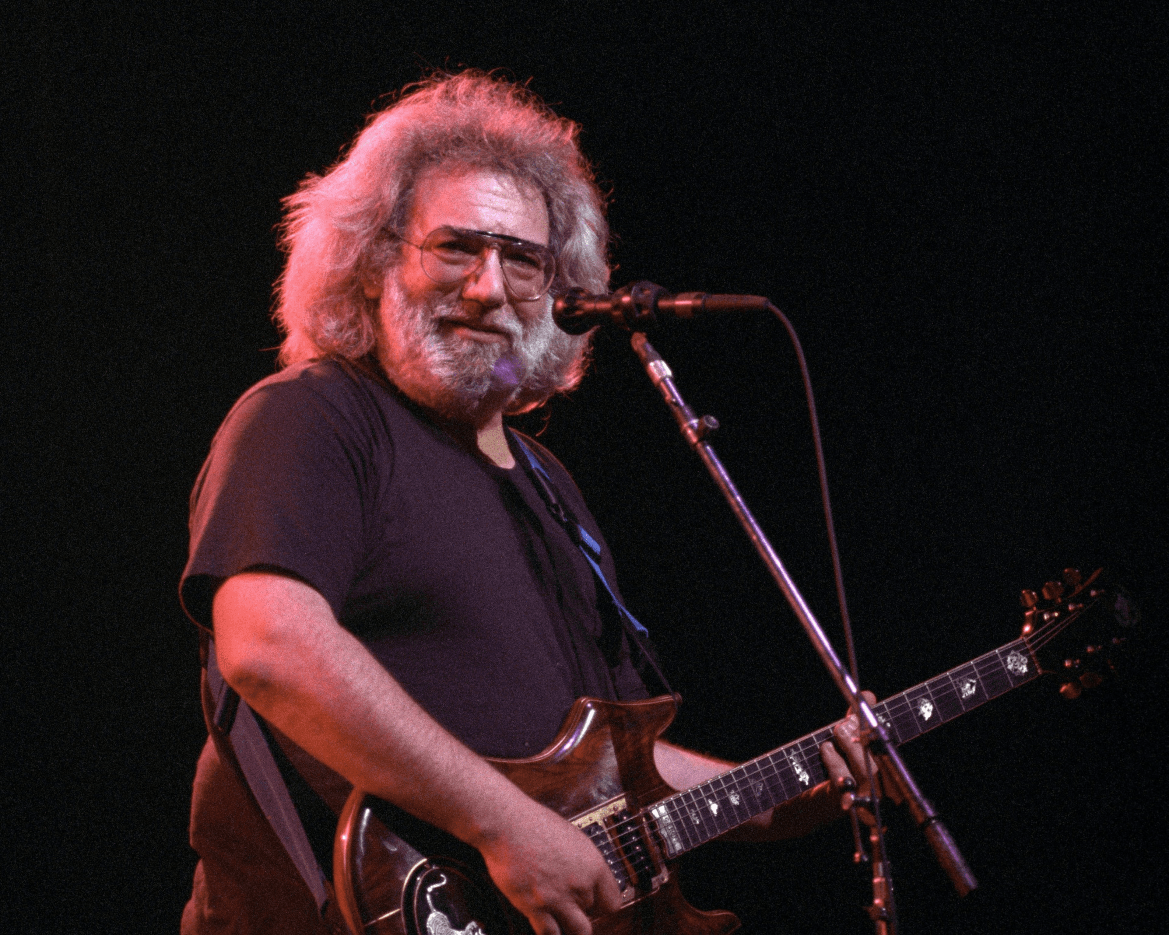 A bearded man with glasses plays an electric guitar on stage, smiling towards the audience. The background is dark, highlighting him under warm lighting.