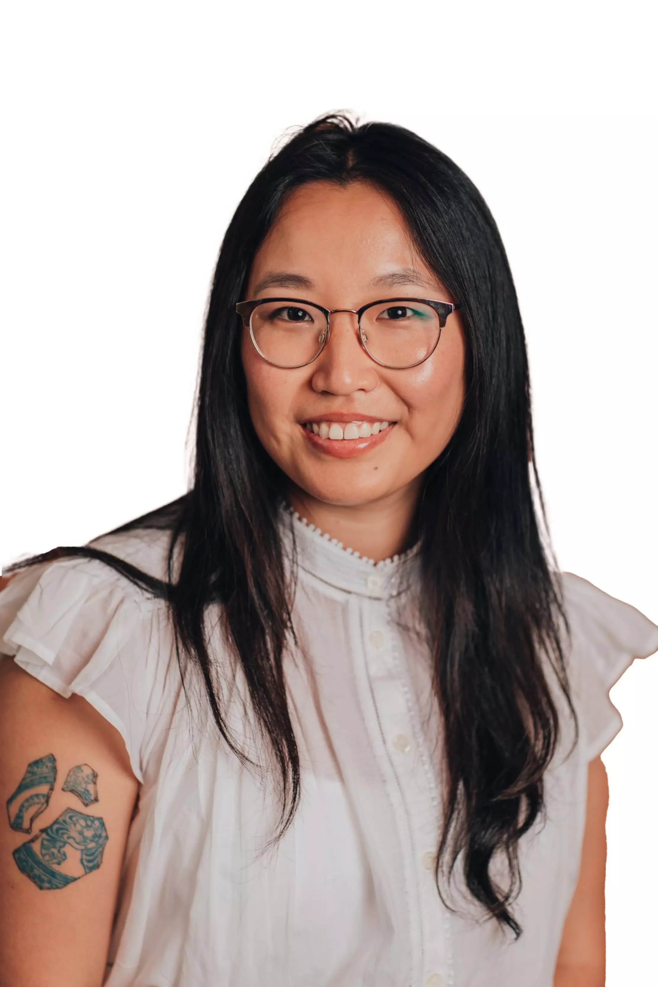 A smiling woman with long black hair and glasses wears a white blouse with ruffled sleeves and has a blue tattoo on her upper left arm.
