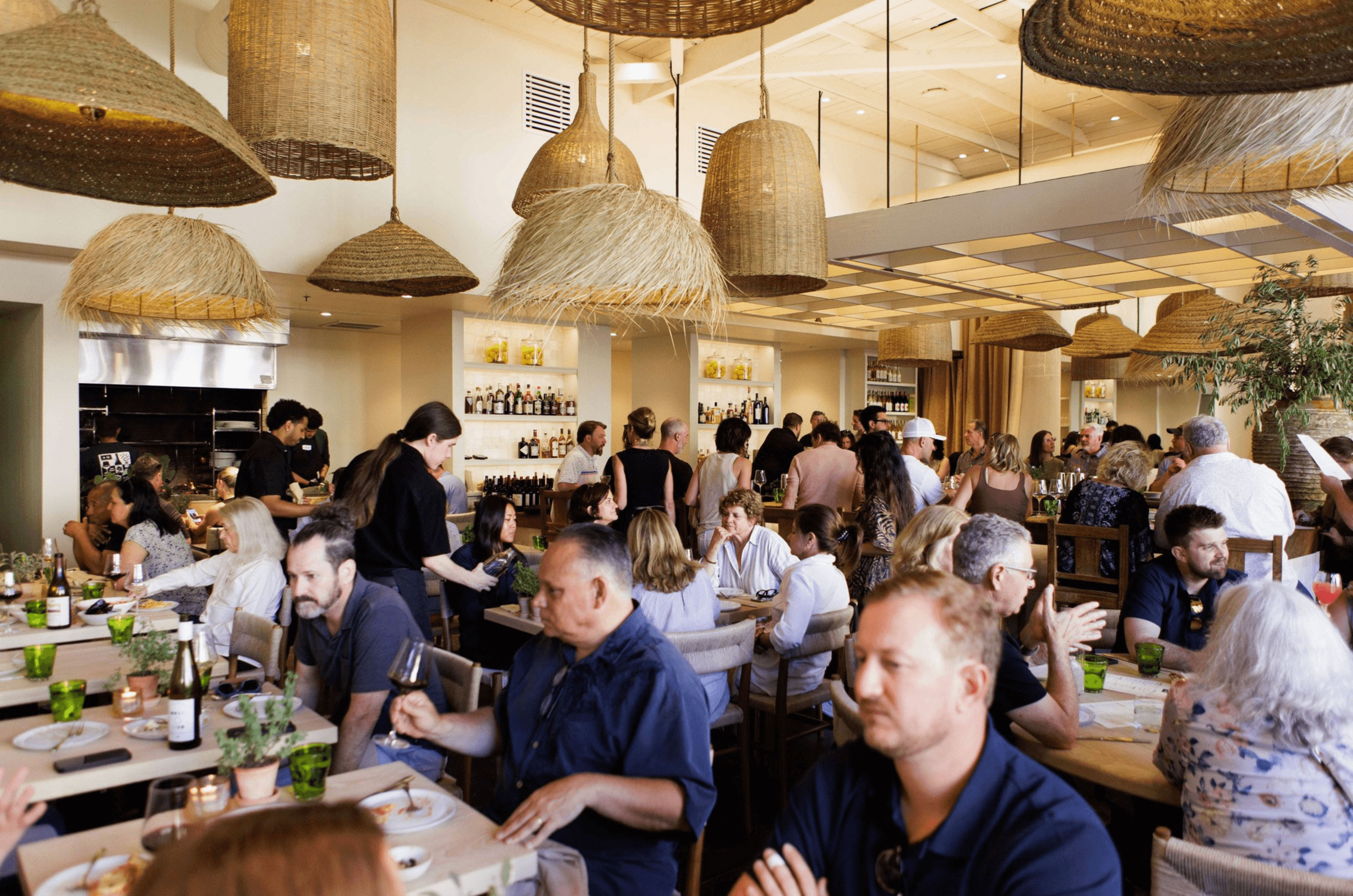 The image shows a bustling restaurant with people dining and chatting at tables. Woven basket-like lights hang from the ceiling, and a bar is visible in the background.