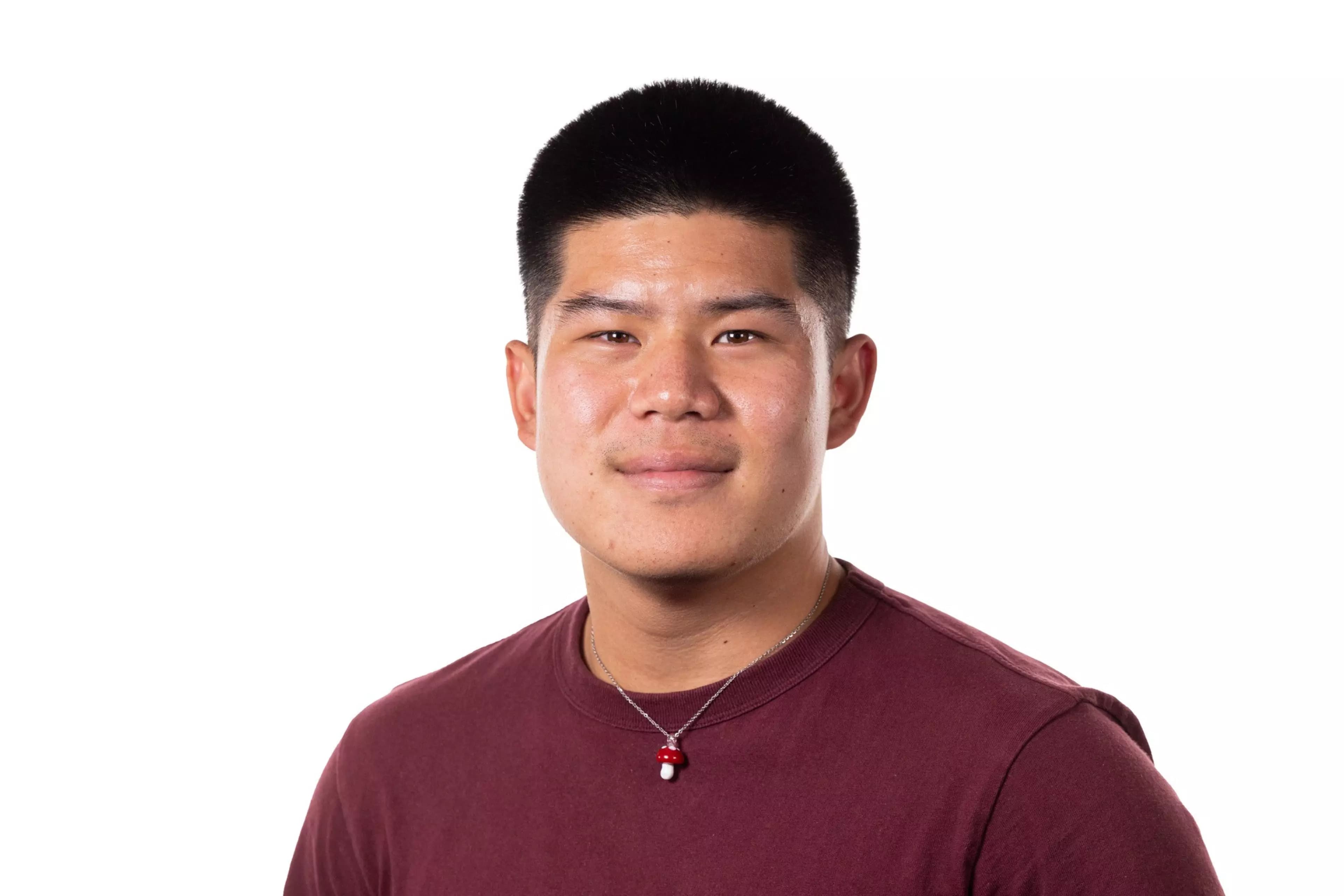 A young man with short black hair wears a maroon shirt and a necklace with a small red and white mushroom pendant, smiling slightly against a white background.