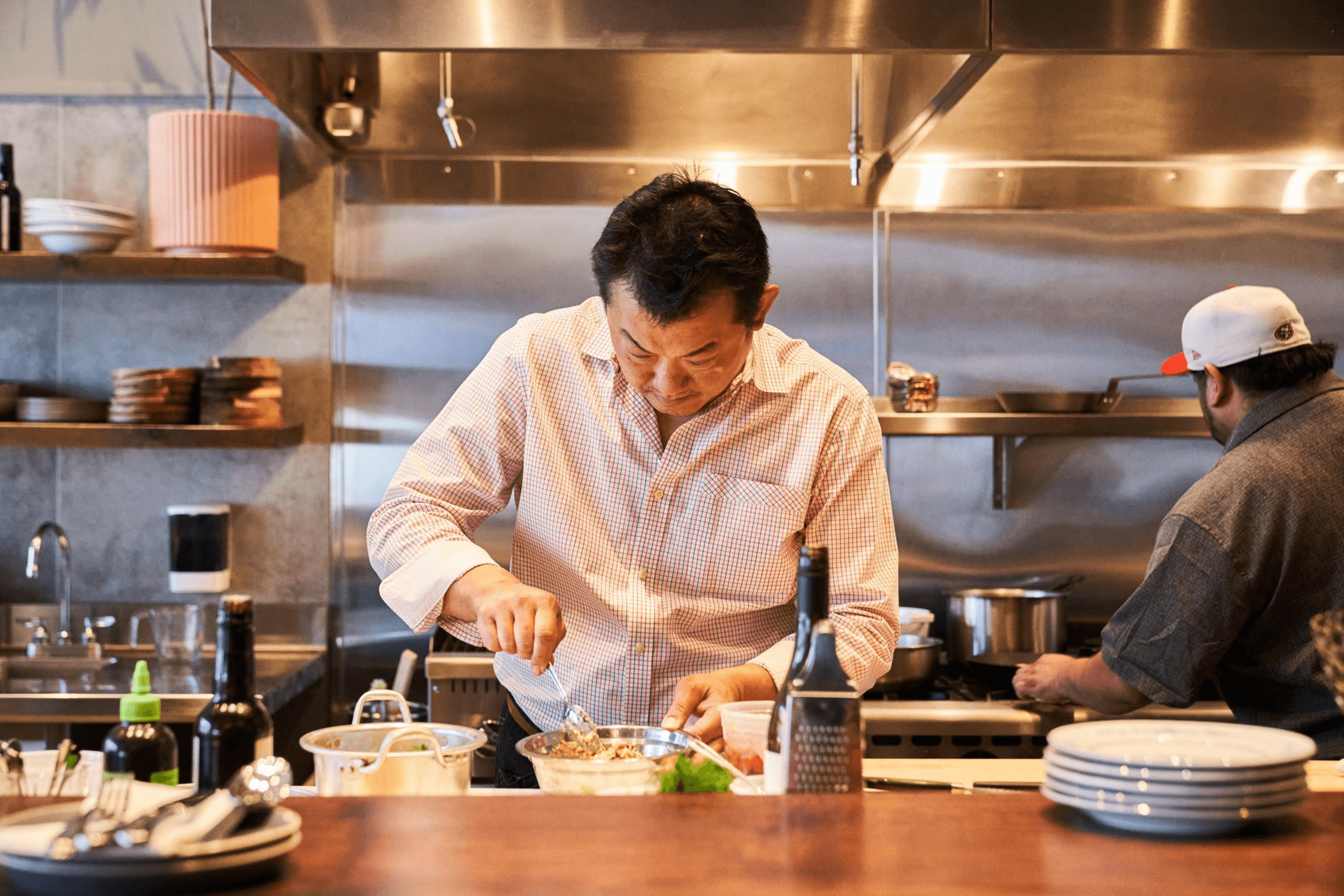 Two chefs are in a professional kitchen. One is focusing on mixing ingredients in a bowl, while the other is cooking at the stove. Shelves with dishes are visible.