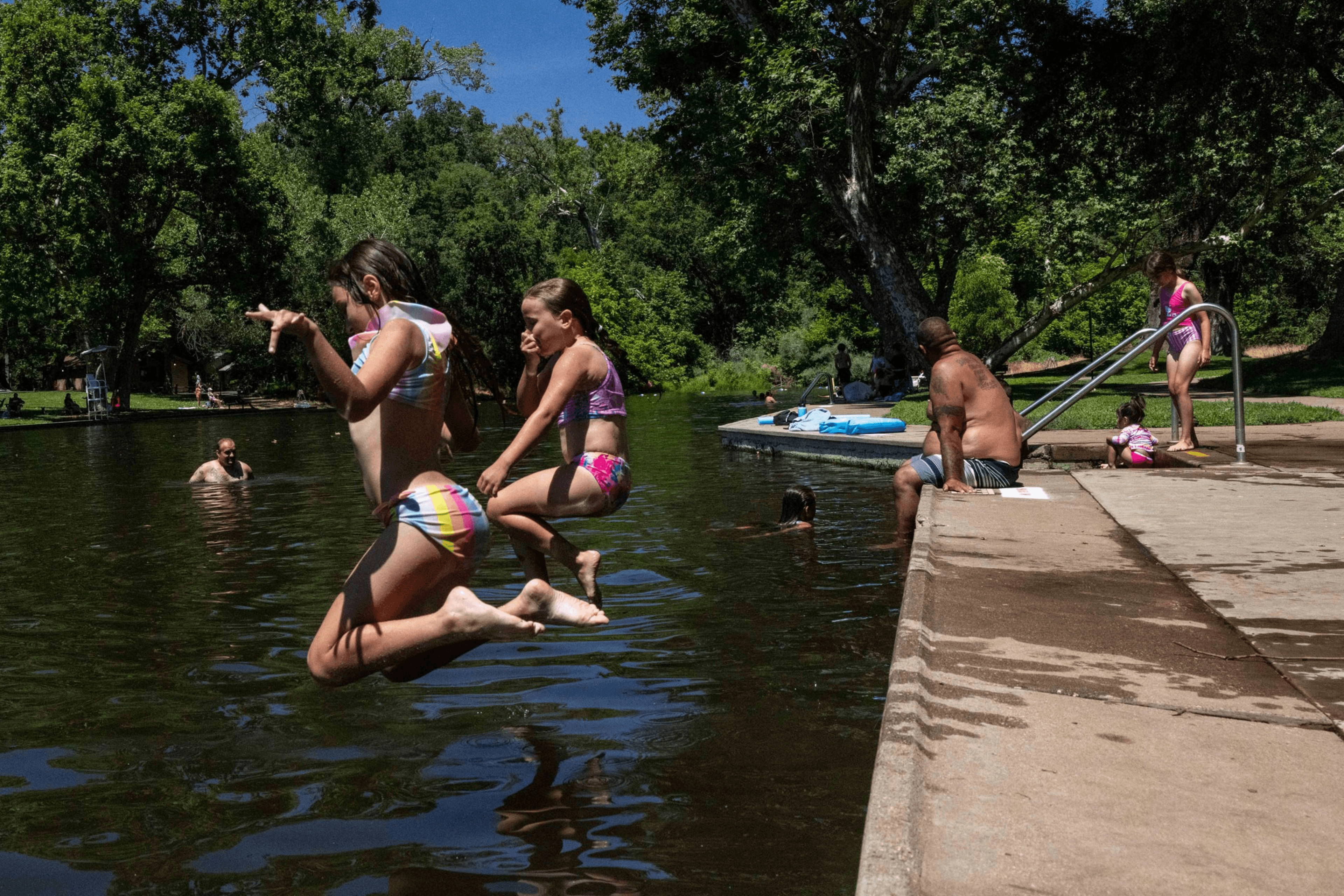 Two girls jump into a lake; nearby, a man and two kids sit on a concrete edge. Trees and other people enjoying the water are visible in the background.