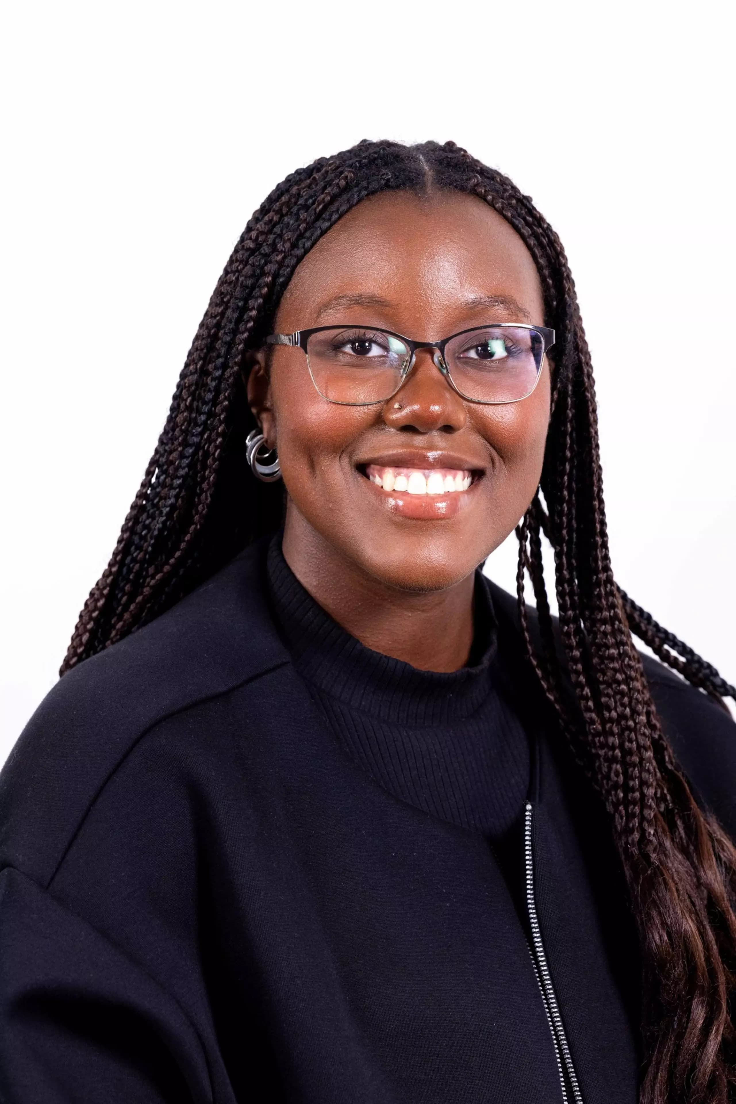 A smiling woman with braided hair, glasses, and hoop earrings wears a black top against a plain white background.