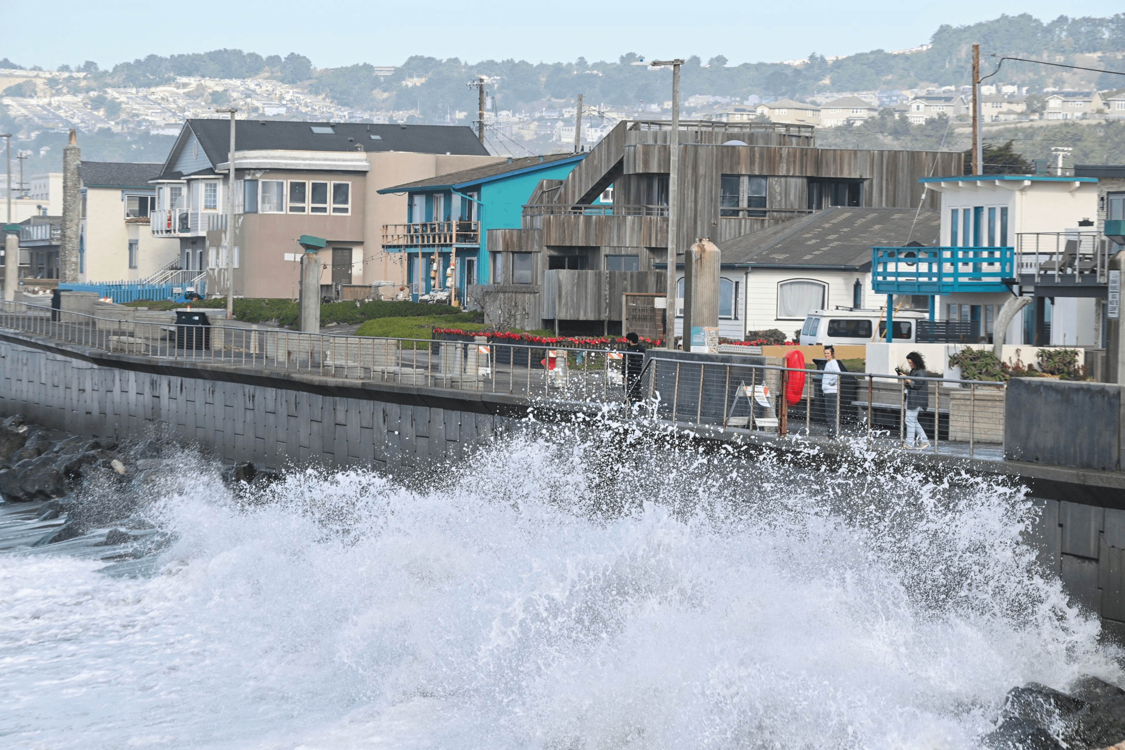 Waves crash against a seawall in front of colorful houses, while people walk along a pathway beside the sea. Hills and more buildings are visible in the background.