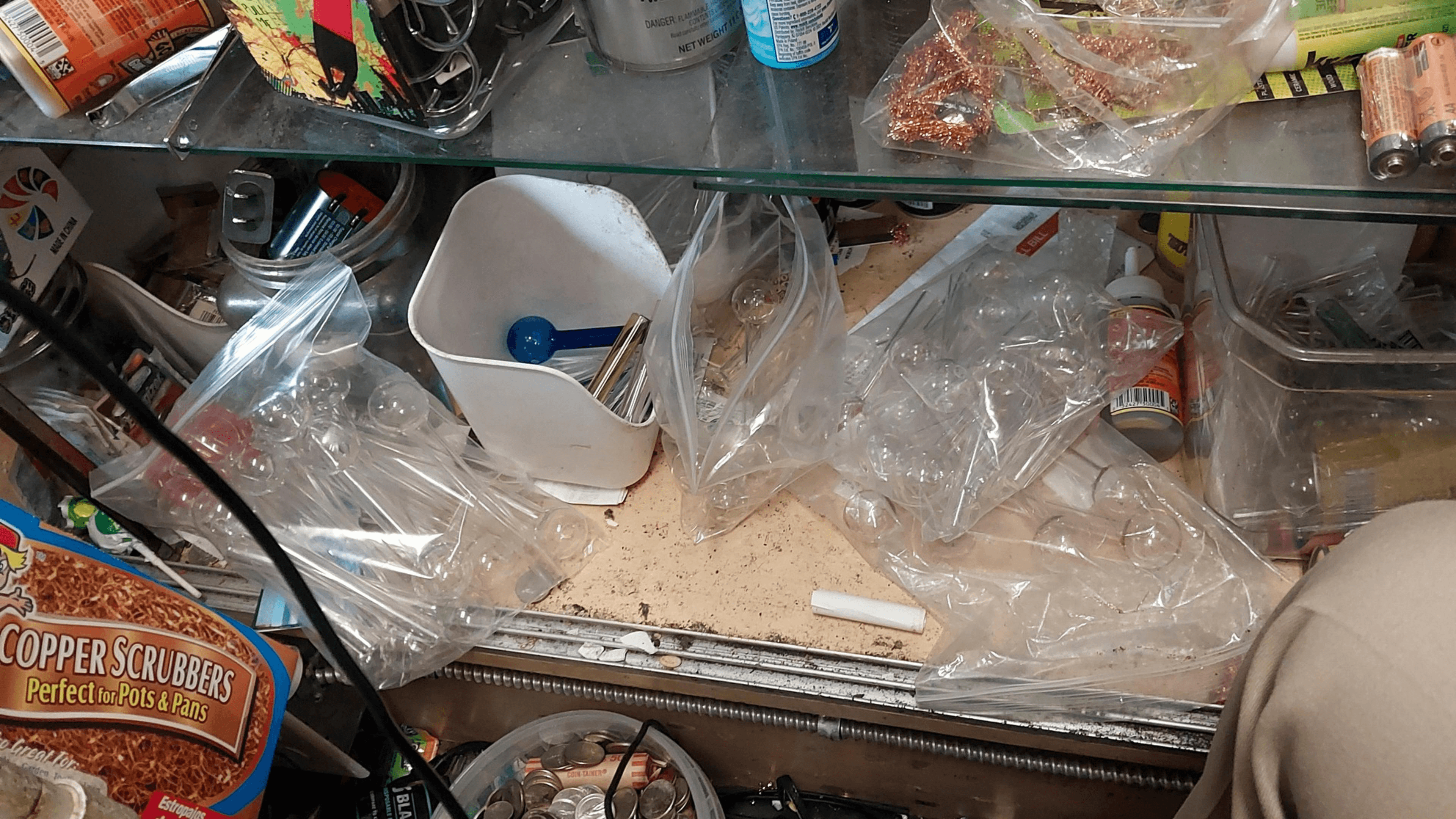 The image shows a cluttered shelf with various items, including batteries, copper scrubbers, plastic bags, and a container with a blue scoop.
