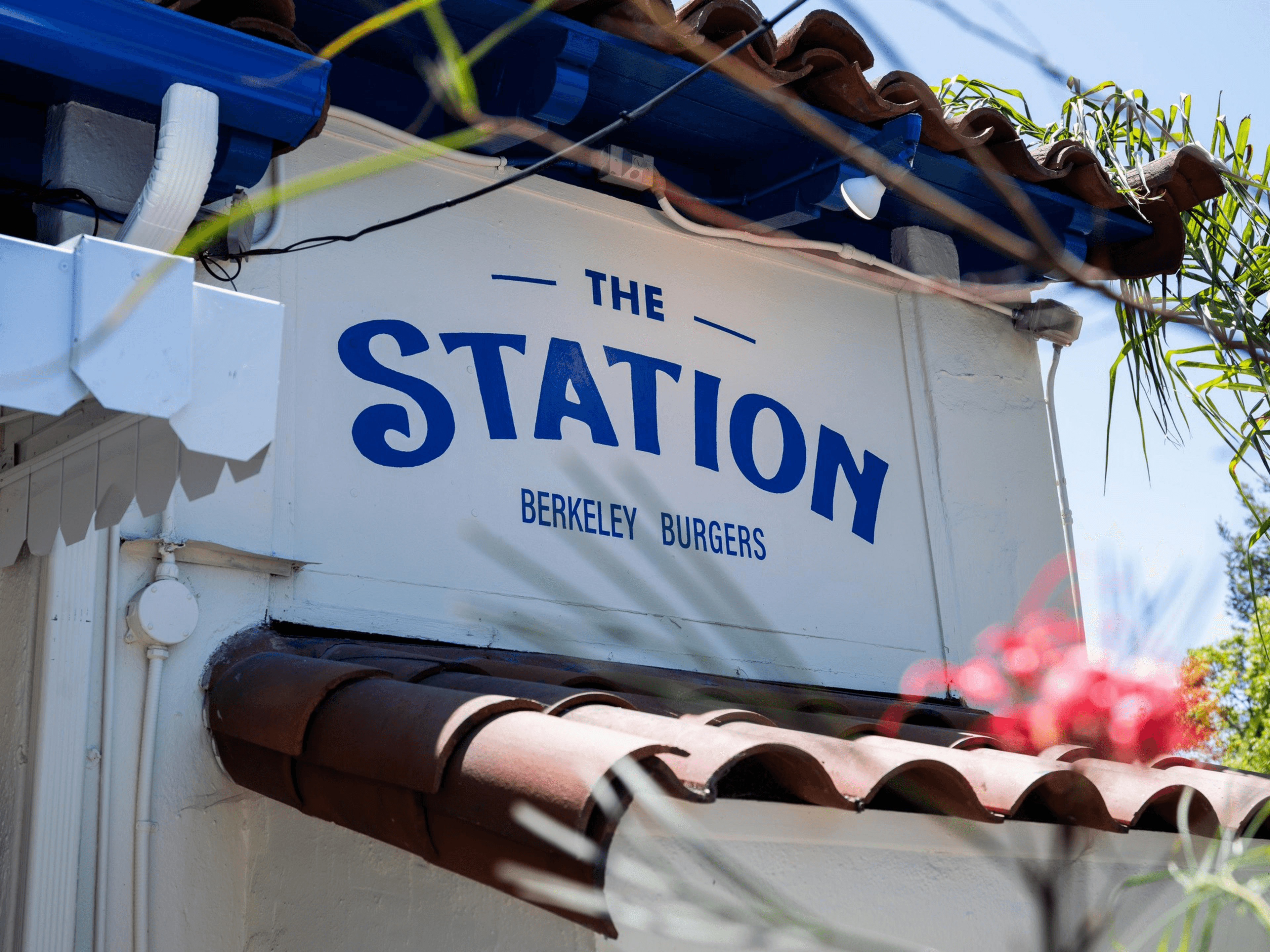 The image shows a sign for "The Station" with "Berkeley Burgers" underneath. It's set on a building with a tiled roof and surrounded by greenery.