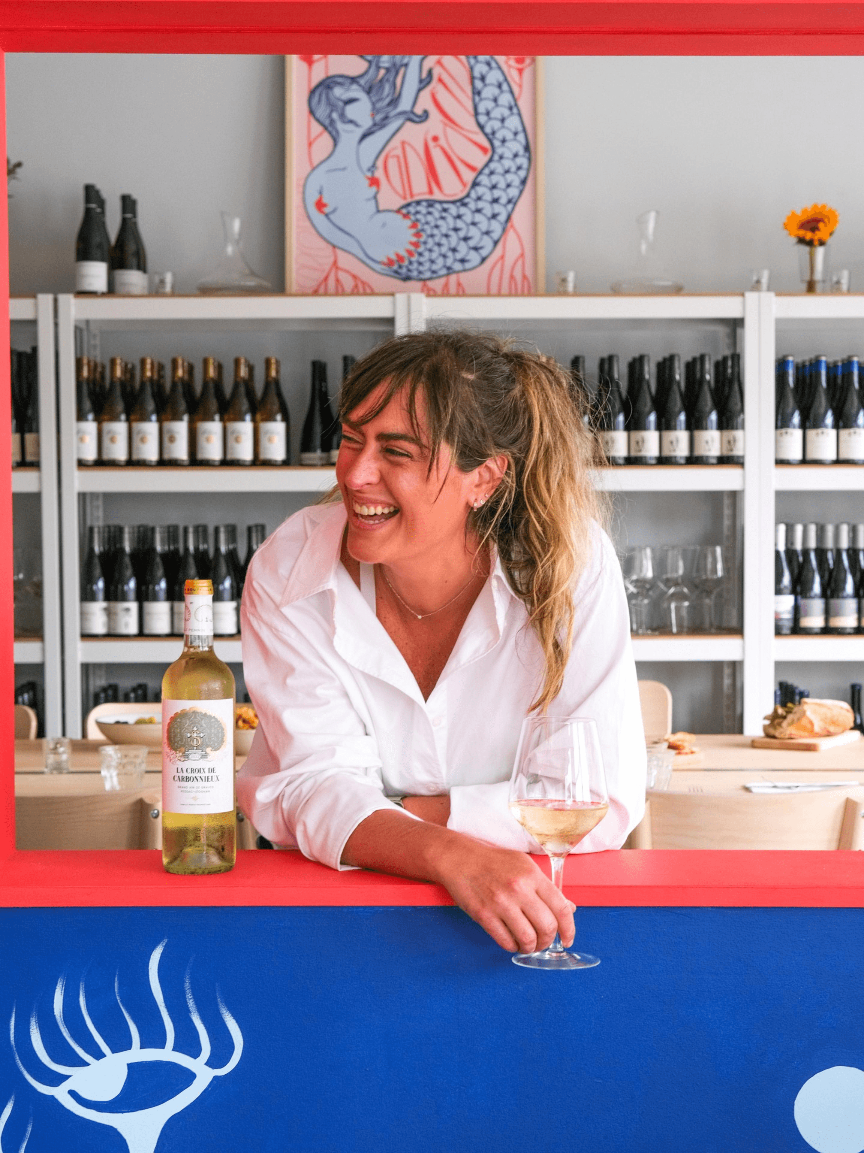 A smiling woman in a white shirt holds a glass of white wine, leaning on a counter with a blue and red backdrop. Shelves of wine bottles and a mermaid painting are behind her.