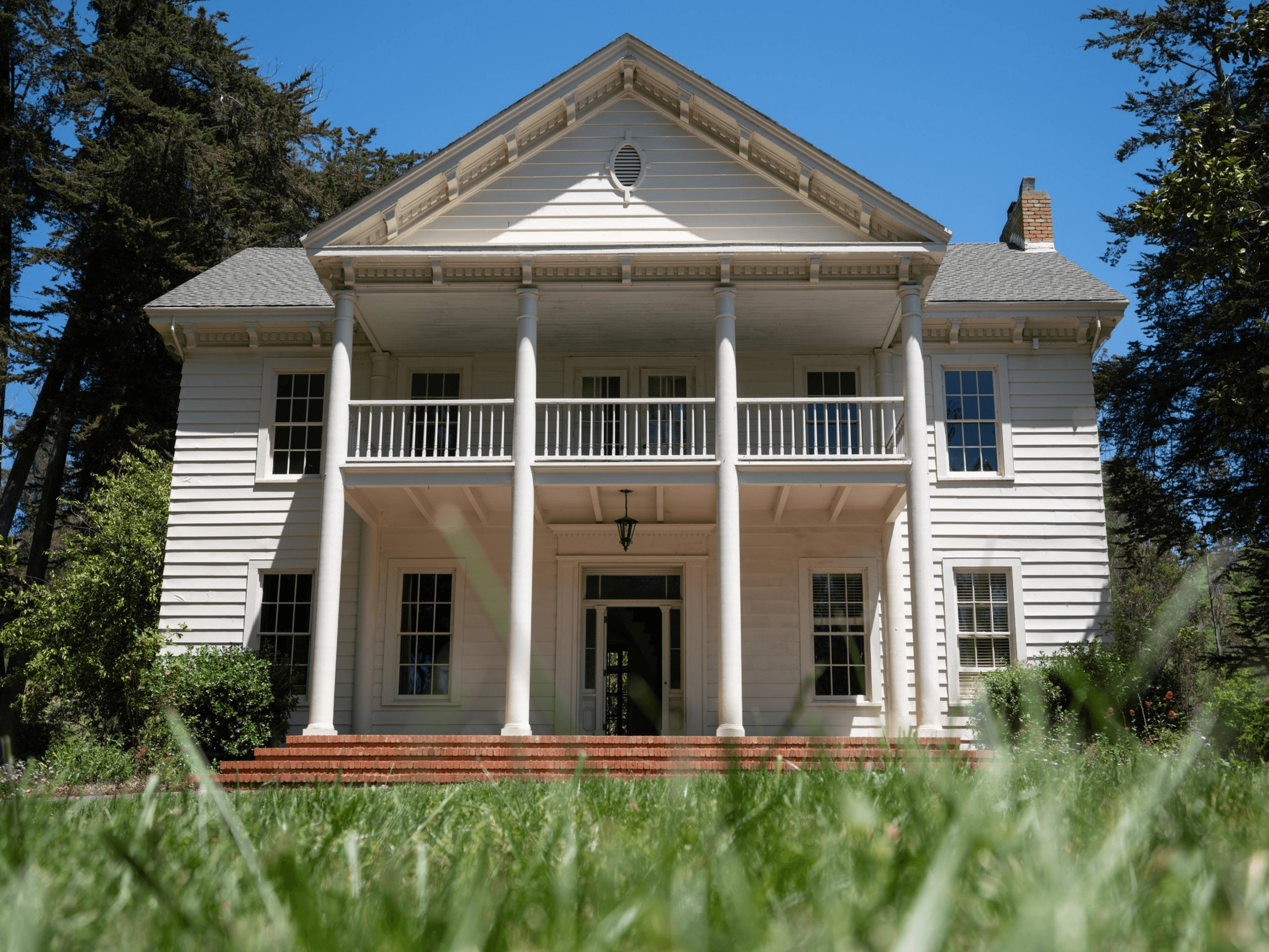 A two-story white house with tall pillars stands against a clear blue sky, surrounded by green trees and shrubs, with a brick step entrance in the foreground.