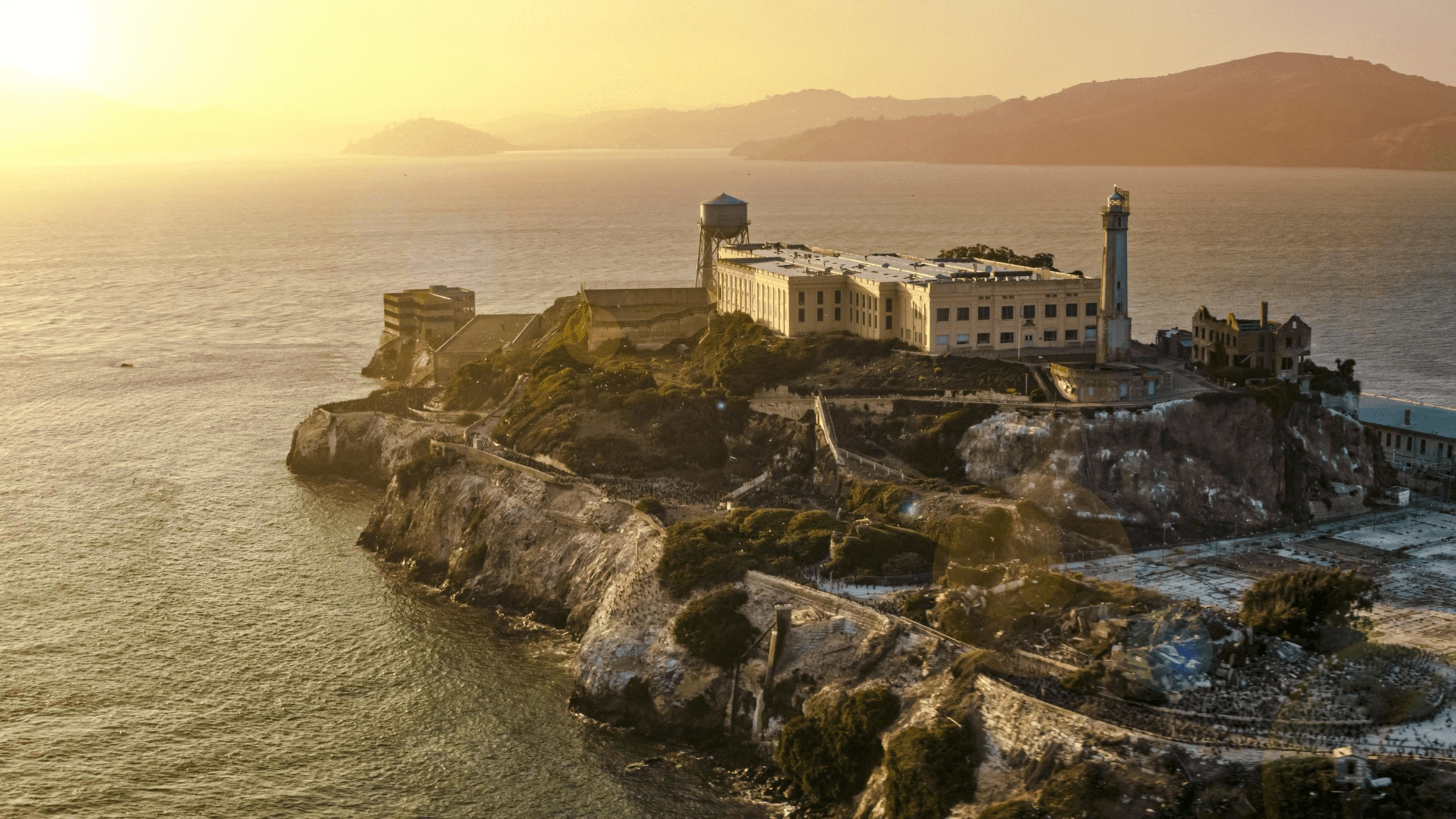 The image shows Alcatraz Island at sunset, with its famous prison building and lighthouse. The island is surrounded by water and a golden sky.