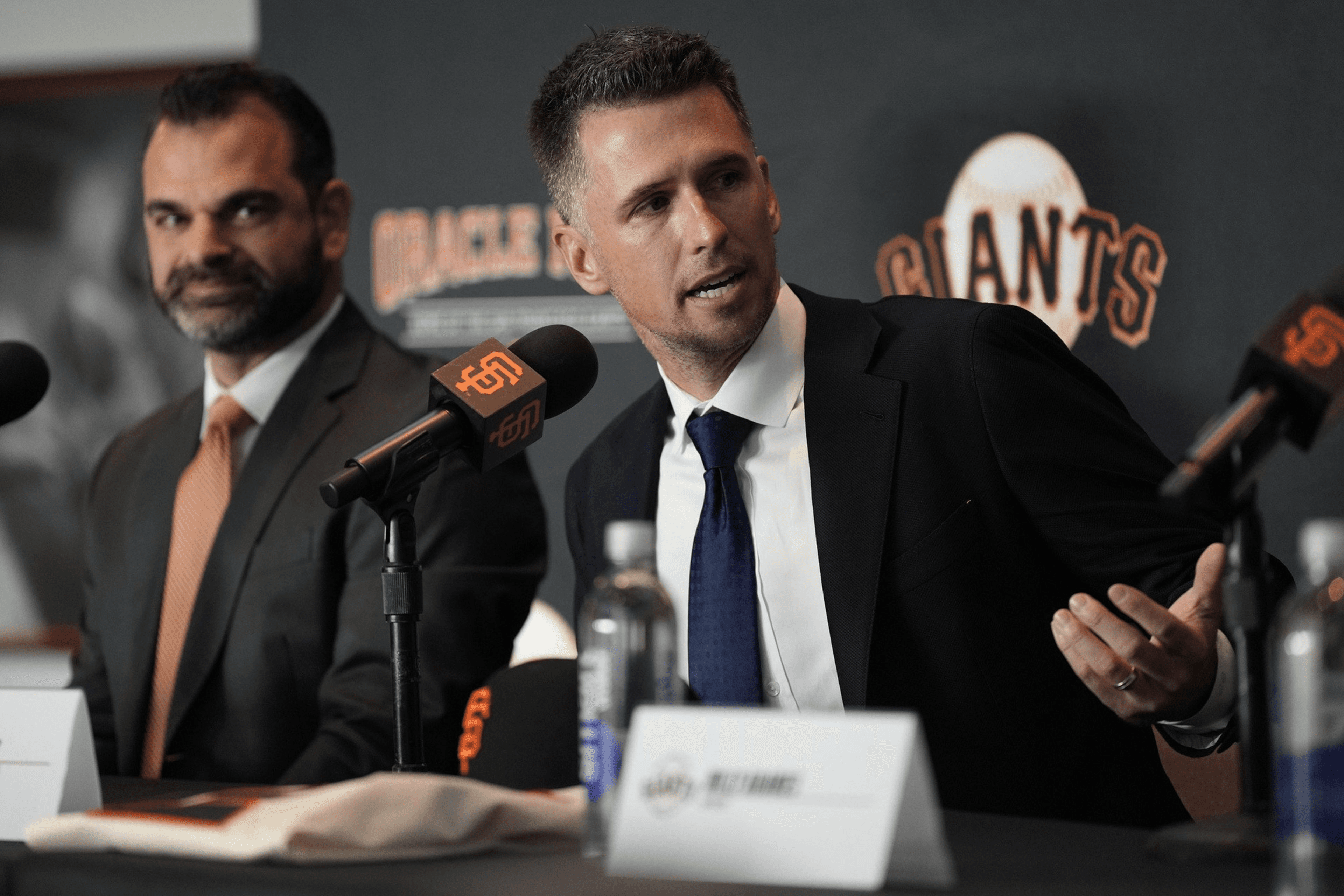 Two men in suits sit at a table with microphones labeled "Giants" in front of them. The man on the right is speaking while gesturing with his hand.