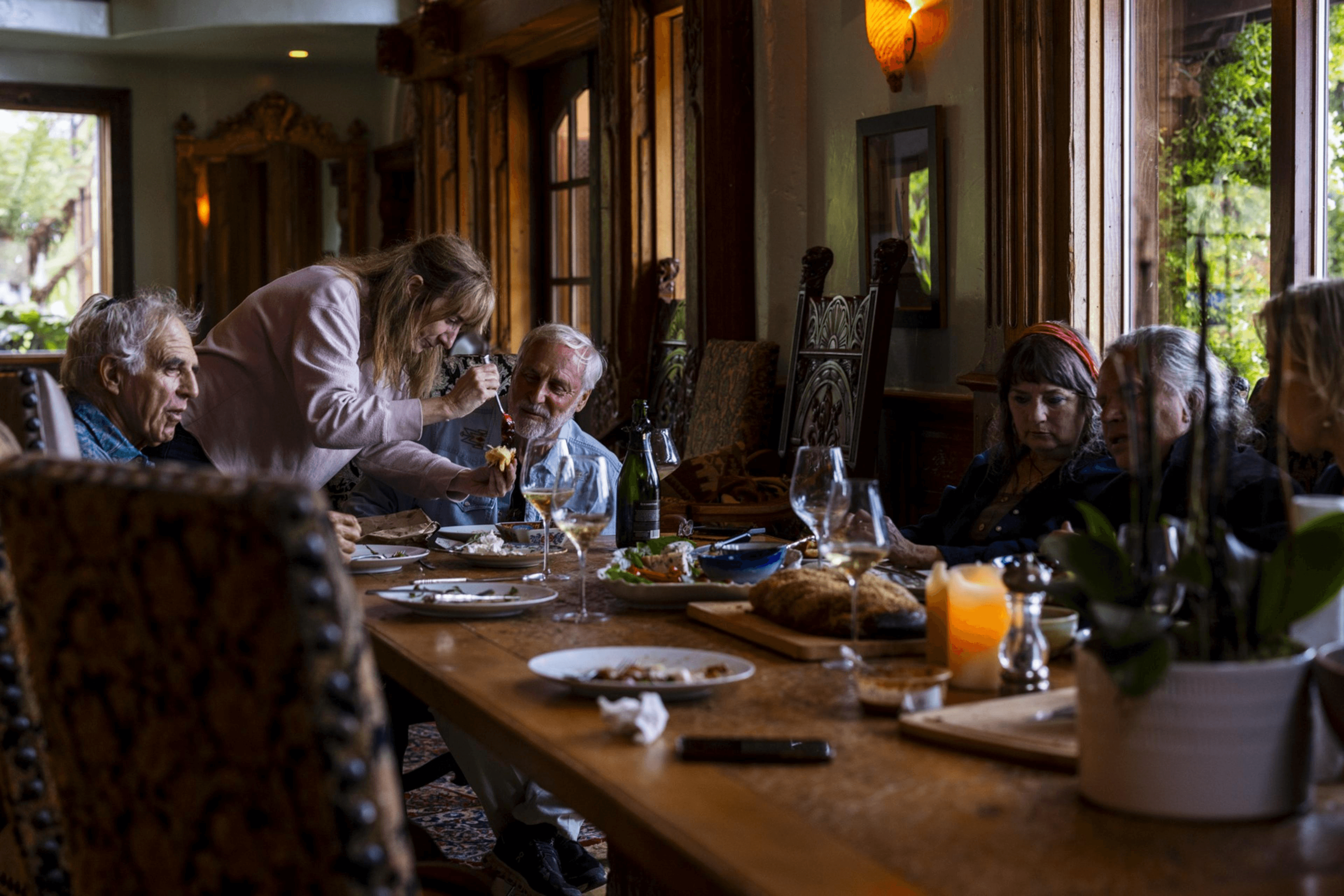 A group of six people is gathered around a dining table indoors. One woman is serving food to an elderly man. The table is set with dishes and a candle.