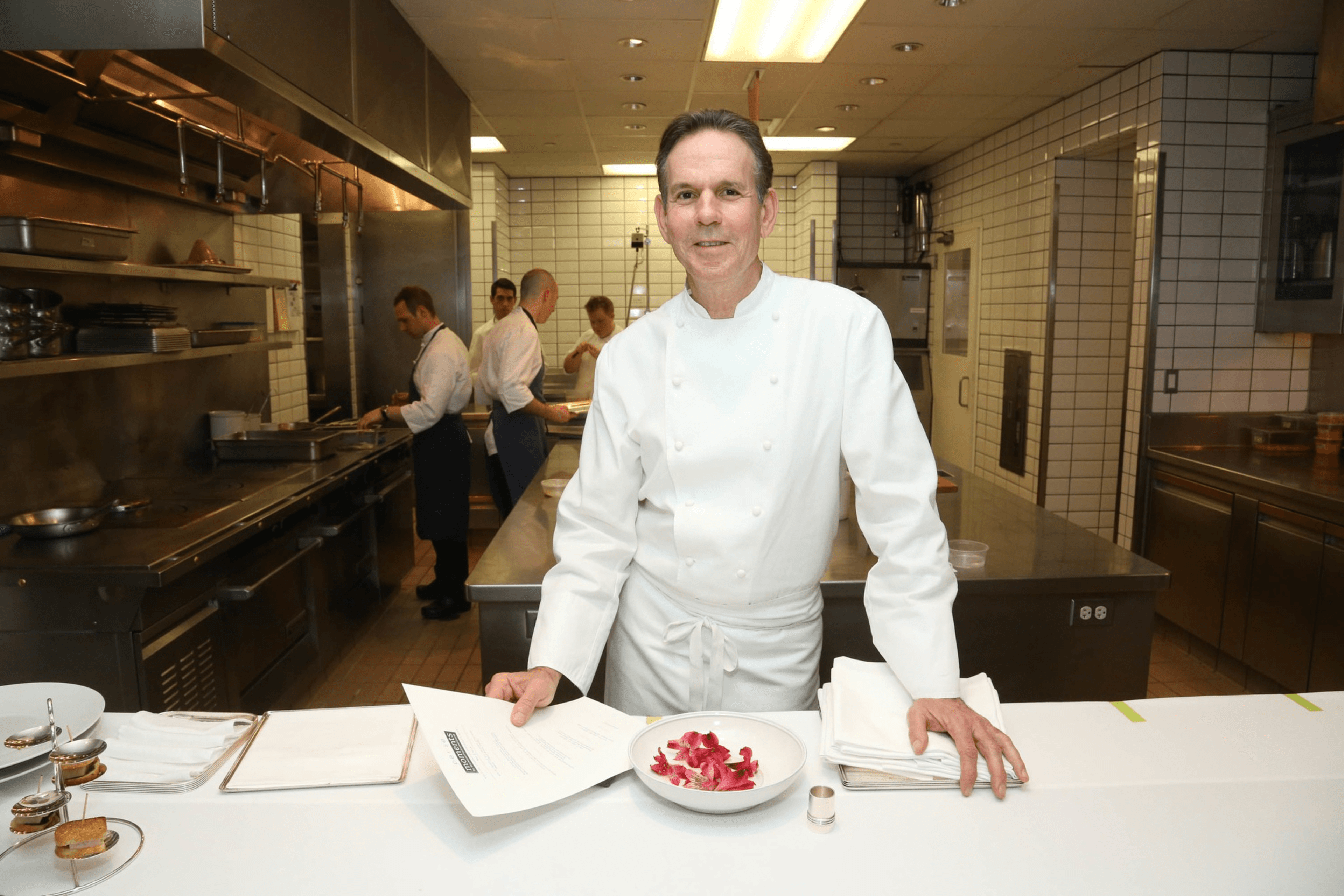 A chef in a white uniform stands in a spotless, tiled kitchen. He holds a paper, with a bowl of red petals nearby. Behind him, others work in the kitchen.