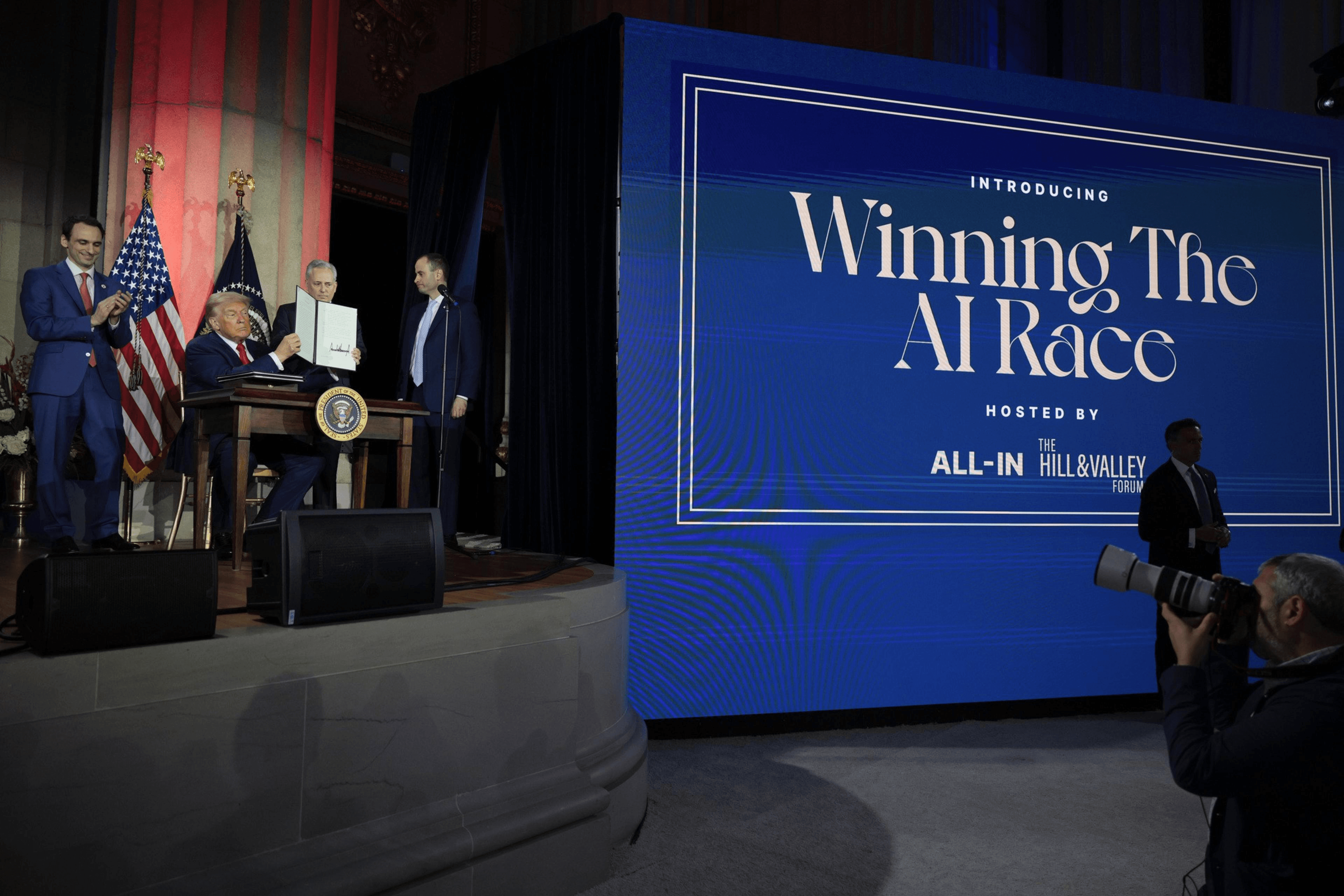 Four men are on stage; one is signing a document at a desk with the presidential seal. A large screen reads "Winning The AI Race" beside them.
