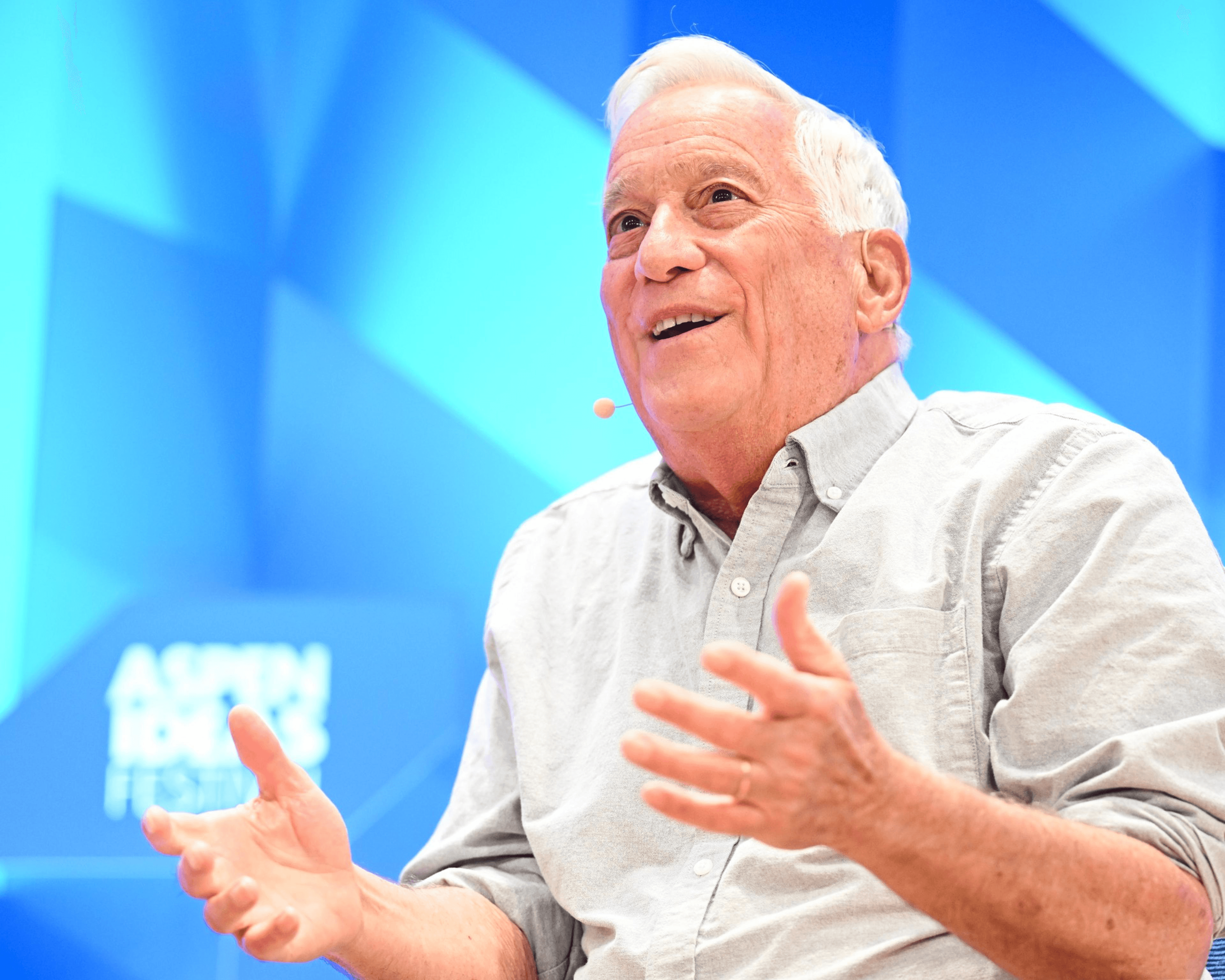 An older man with white hair is speaking animatedly on stage, wearing a light gray shirt. He's gesturing with his hands; a microphone is visible on his cheek.