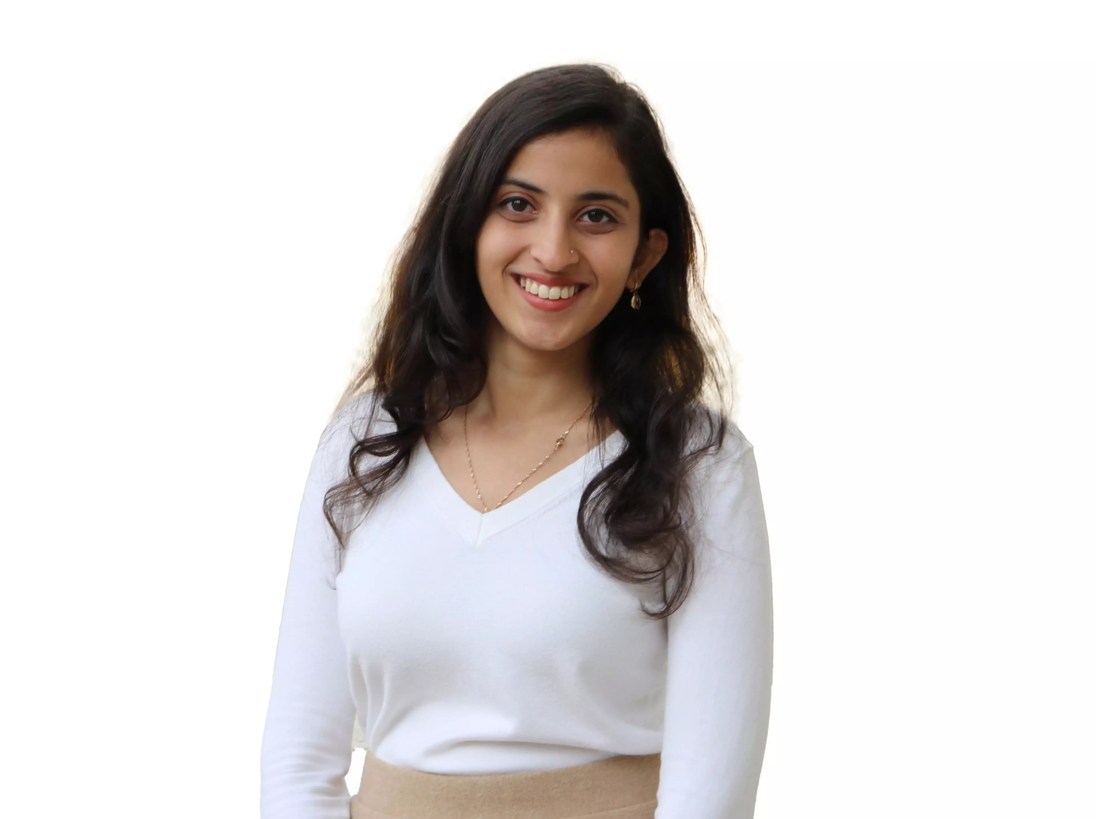 A young woman with long dark hair smiles, wearing a white V-neck sweater, beige skirt, small earrings, and a delicate necklace against a white background.