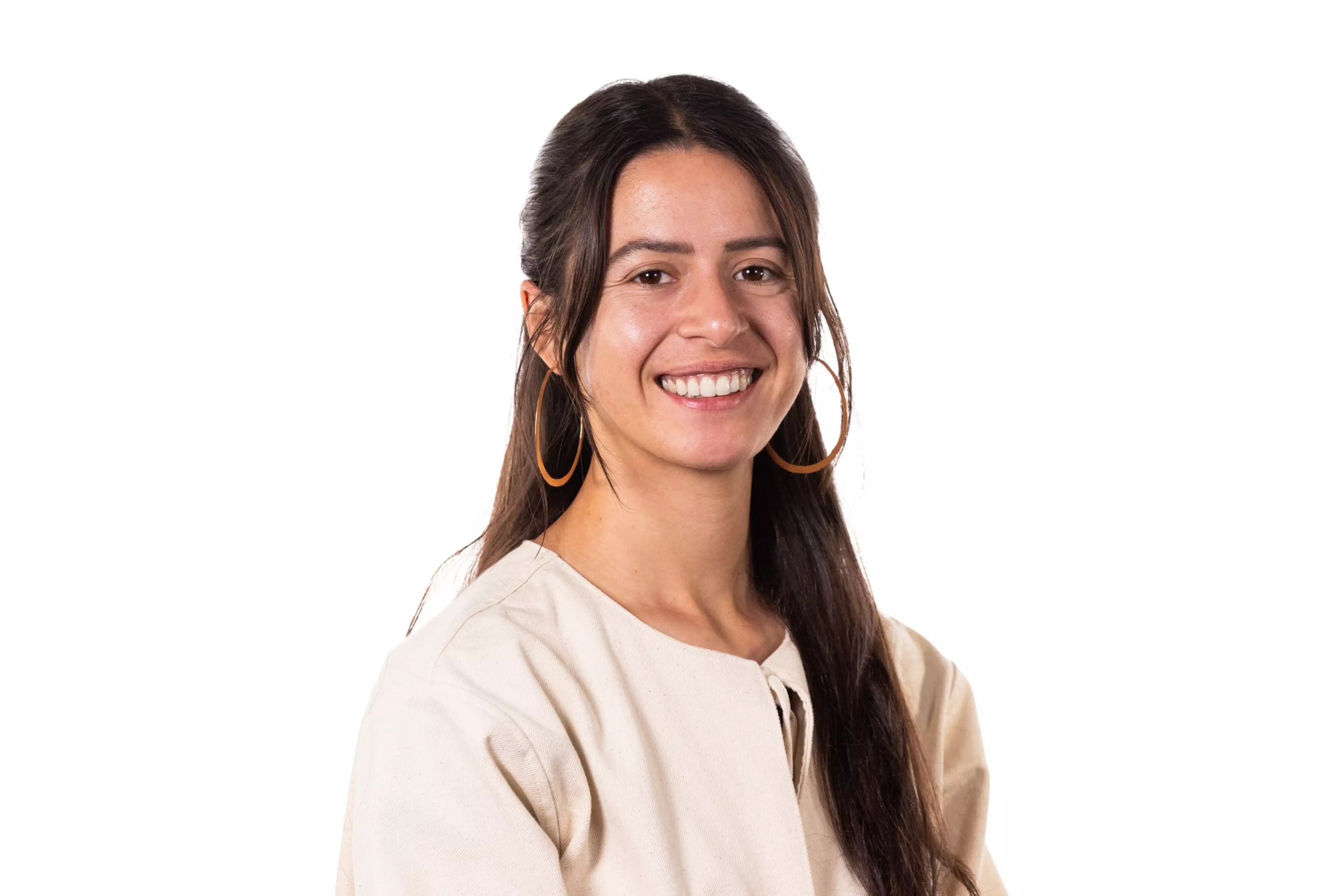 A young woman with long dark hair, wearing large hoop earrings and a light beige top, smiles warmly against a white background.