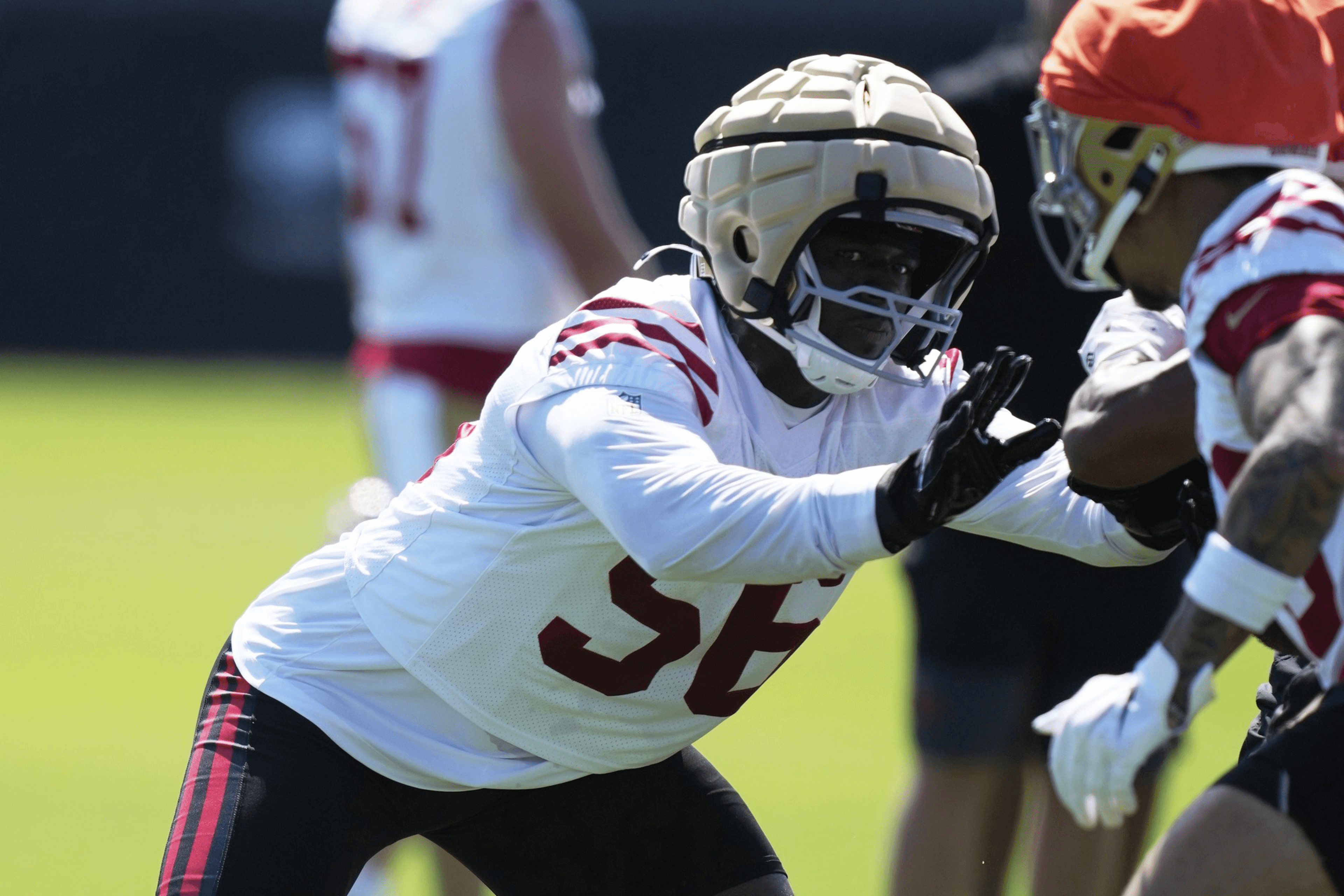 Two American football players in white and red gear are practicing. One wears a padded helmet, and they're engaged in a blocking drill on green grass.