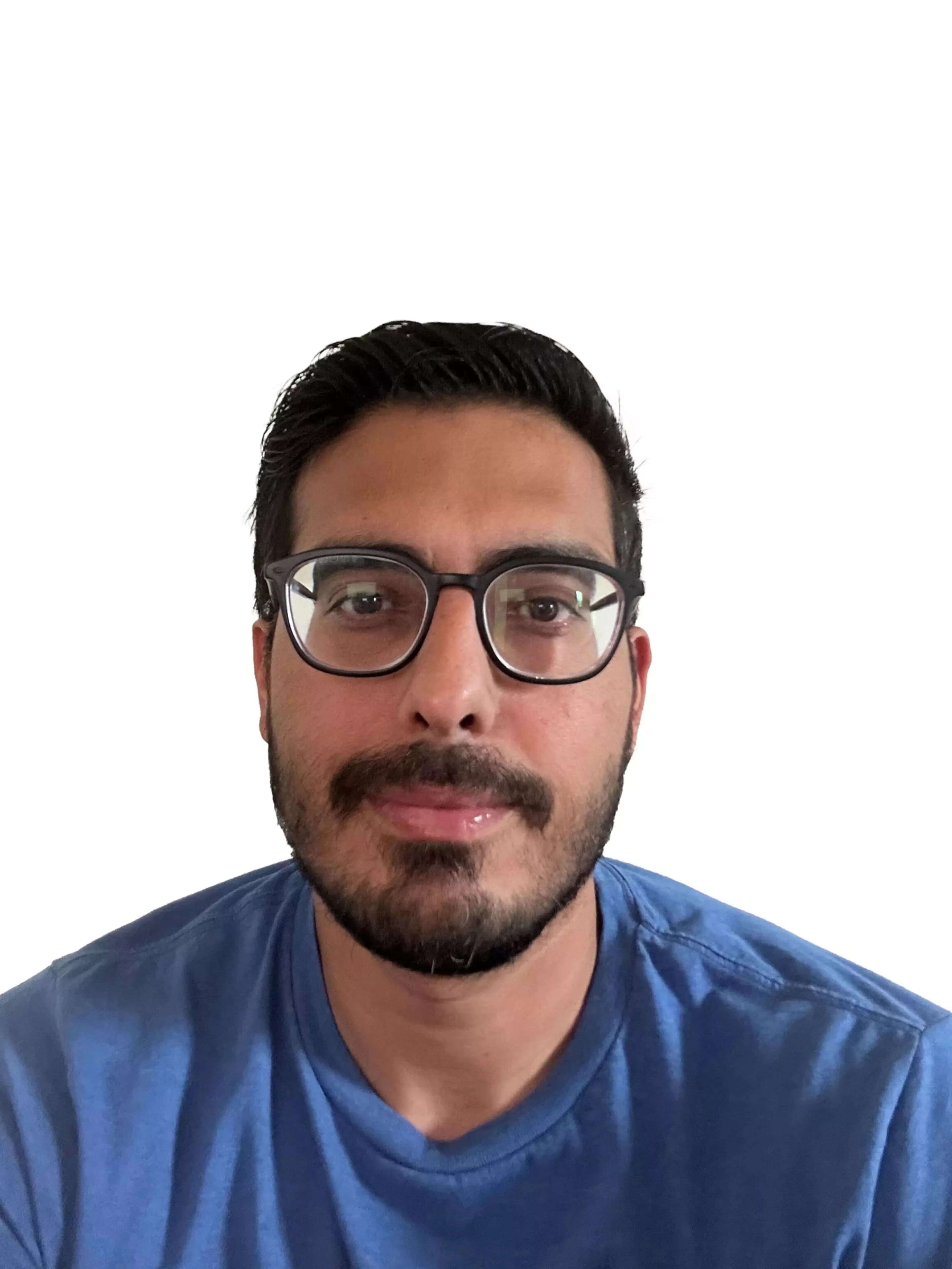 A man with short dark hair, beard, and glasses is wearing a blue shirt, looking straight ahead against a plain white background.