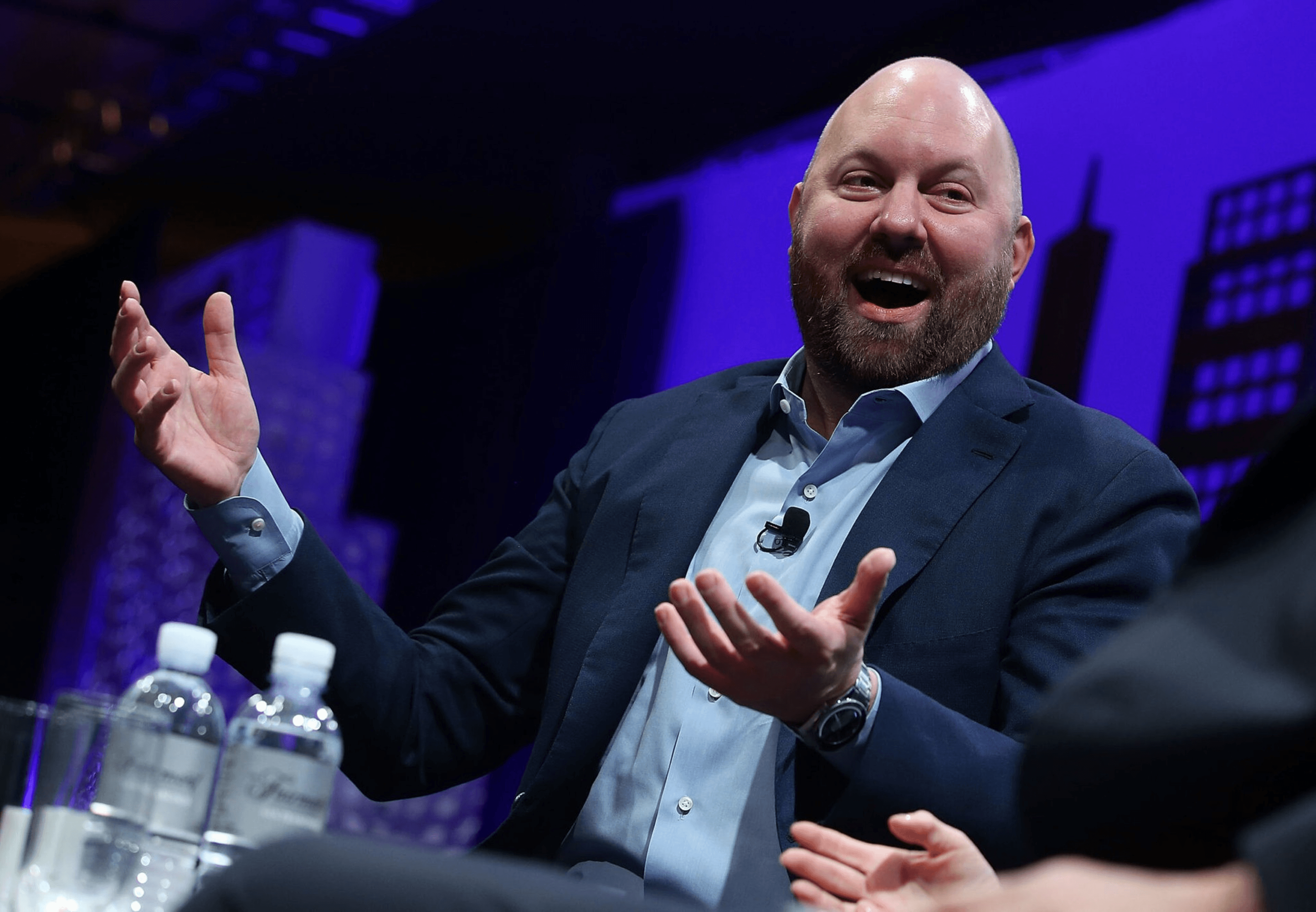 A bald man with a beard, dressed in a suit, is speaking animatedly with a smile, gesturing with his hands. Two bottles of water are in the foreground.